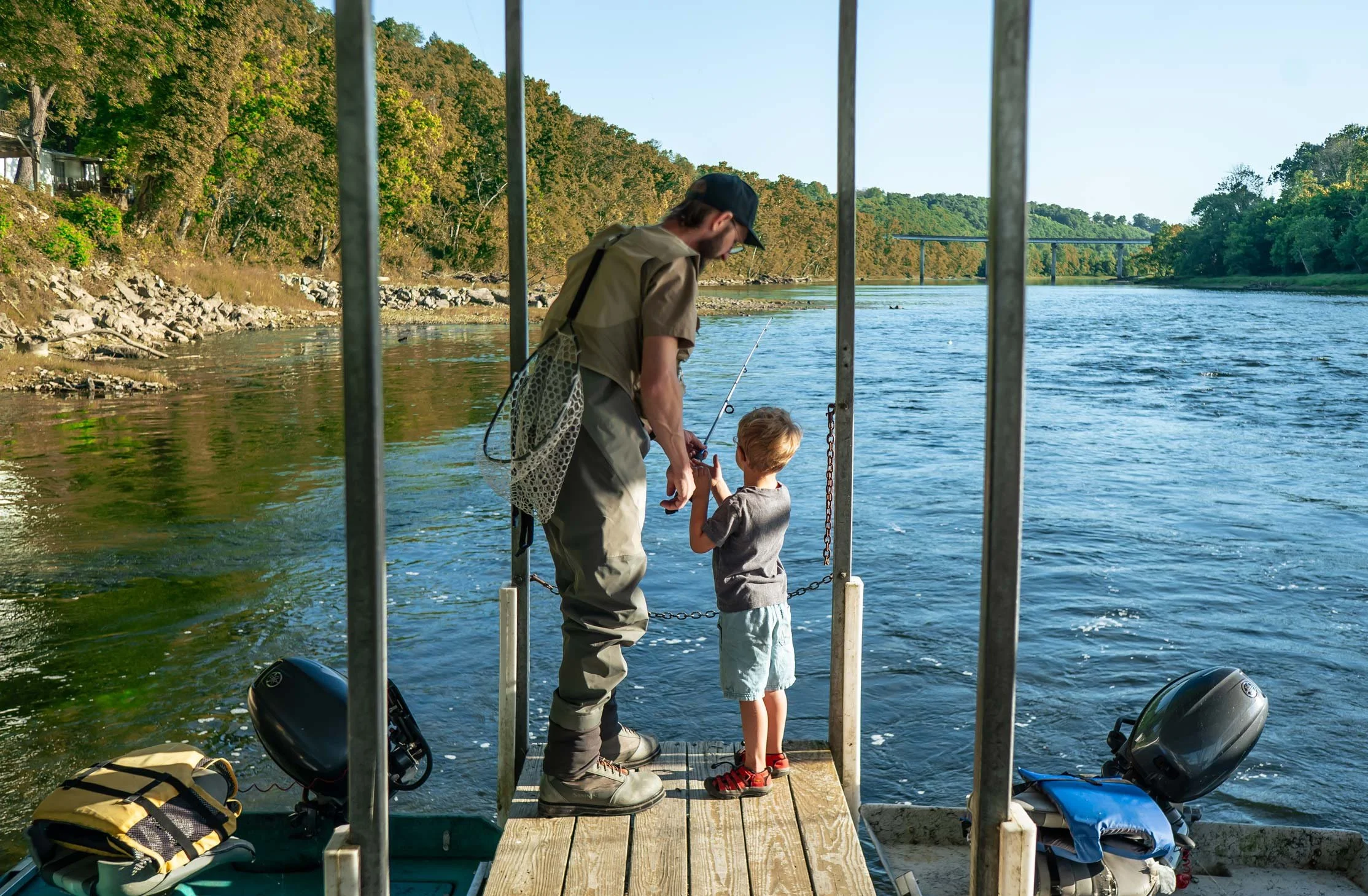 A man and a young boy standing on a wooden dock by the water, fishing. The dock is surrounded by trees and a bridge is visible in the background.