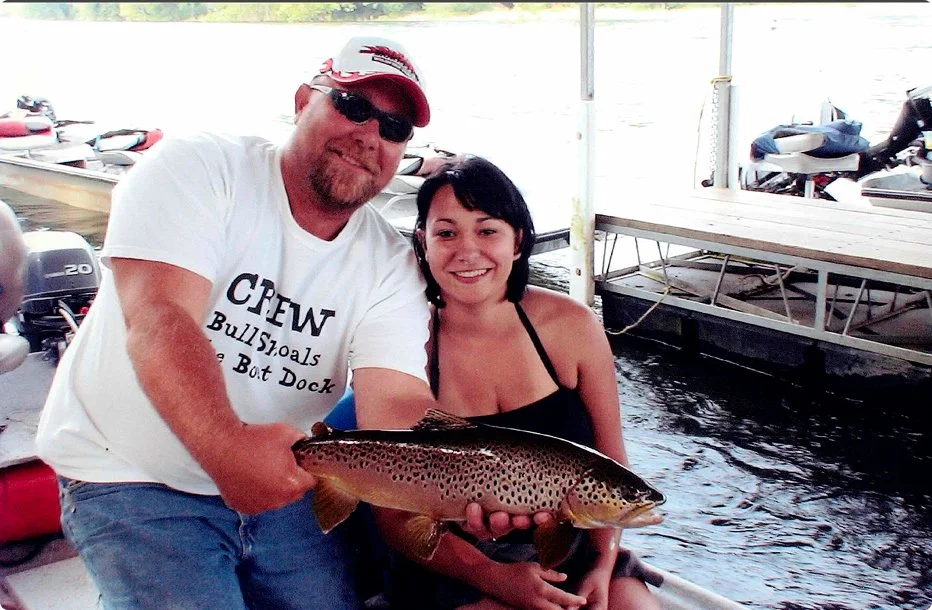 A man and a woman posing with a large fish in a boat docked at a marina, with boats and water in the background.