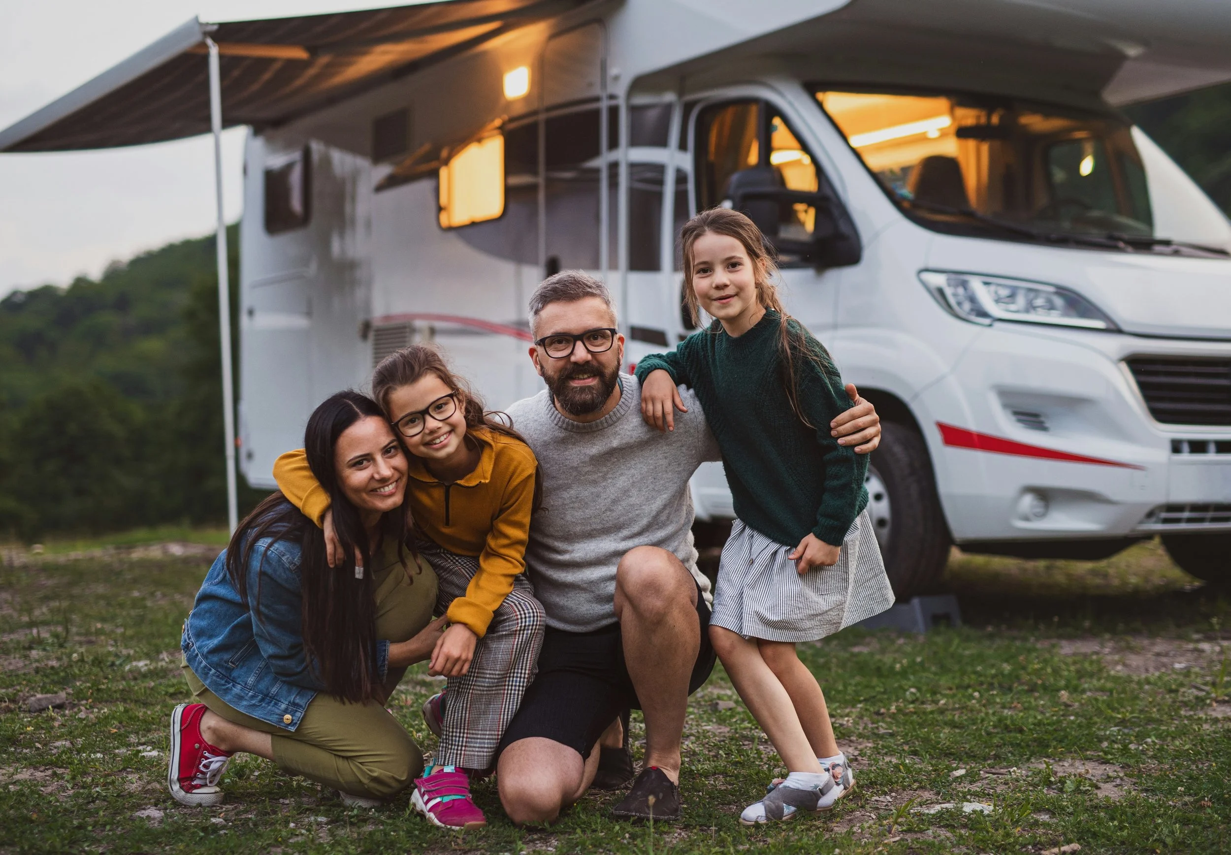 A family of five, including a man, woman, three young girls, is smiling and embracing each other in front of a large motorhome parked outdoors during sunset or early evening.