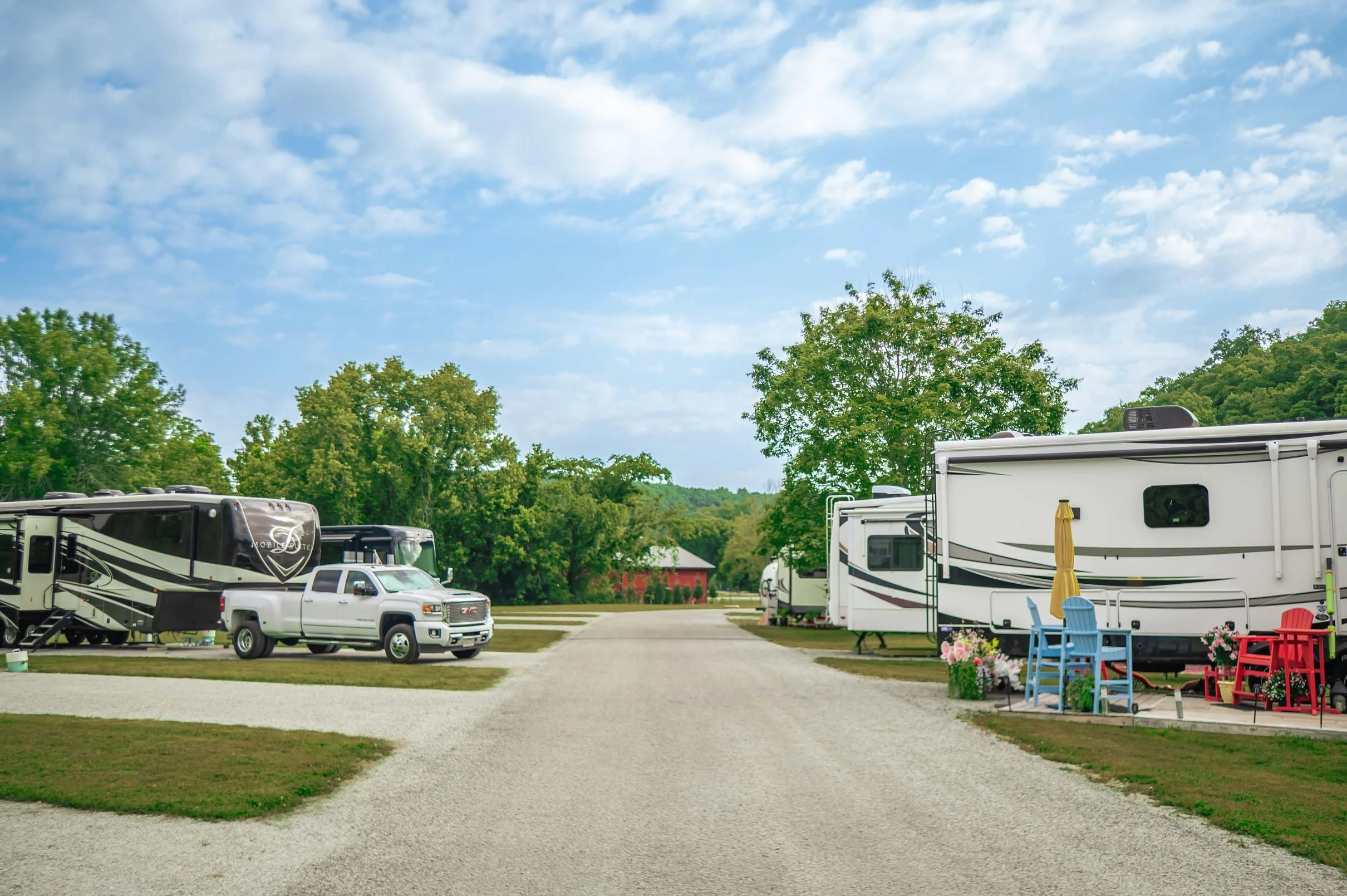 A campground with multiple RVs and trailers parked along a gravel path, green trees in the background, a small red barn, and outdoor furniture with colorful decorations under a partly cloudy sky.