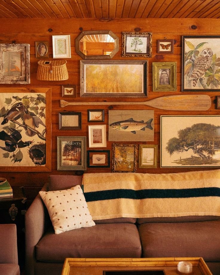 A living room with a brown couch, a cream blanket with black stripe, a white pillow with small star pattern, and a gallery wall of various framed pictures, paintings, and mirrors on a wood-paneled wall.