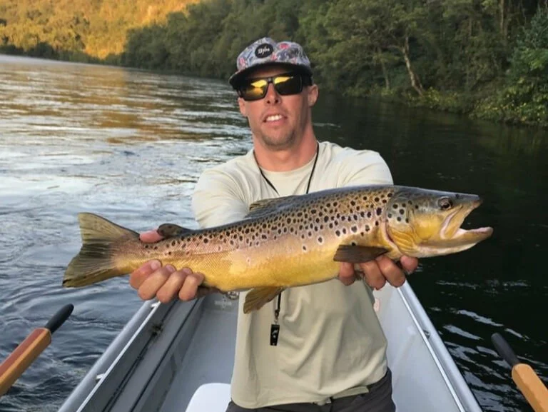 Man in a boat holding a large brown and yellow trout with black spots, on a river with trees in the background.