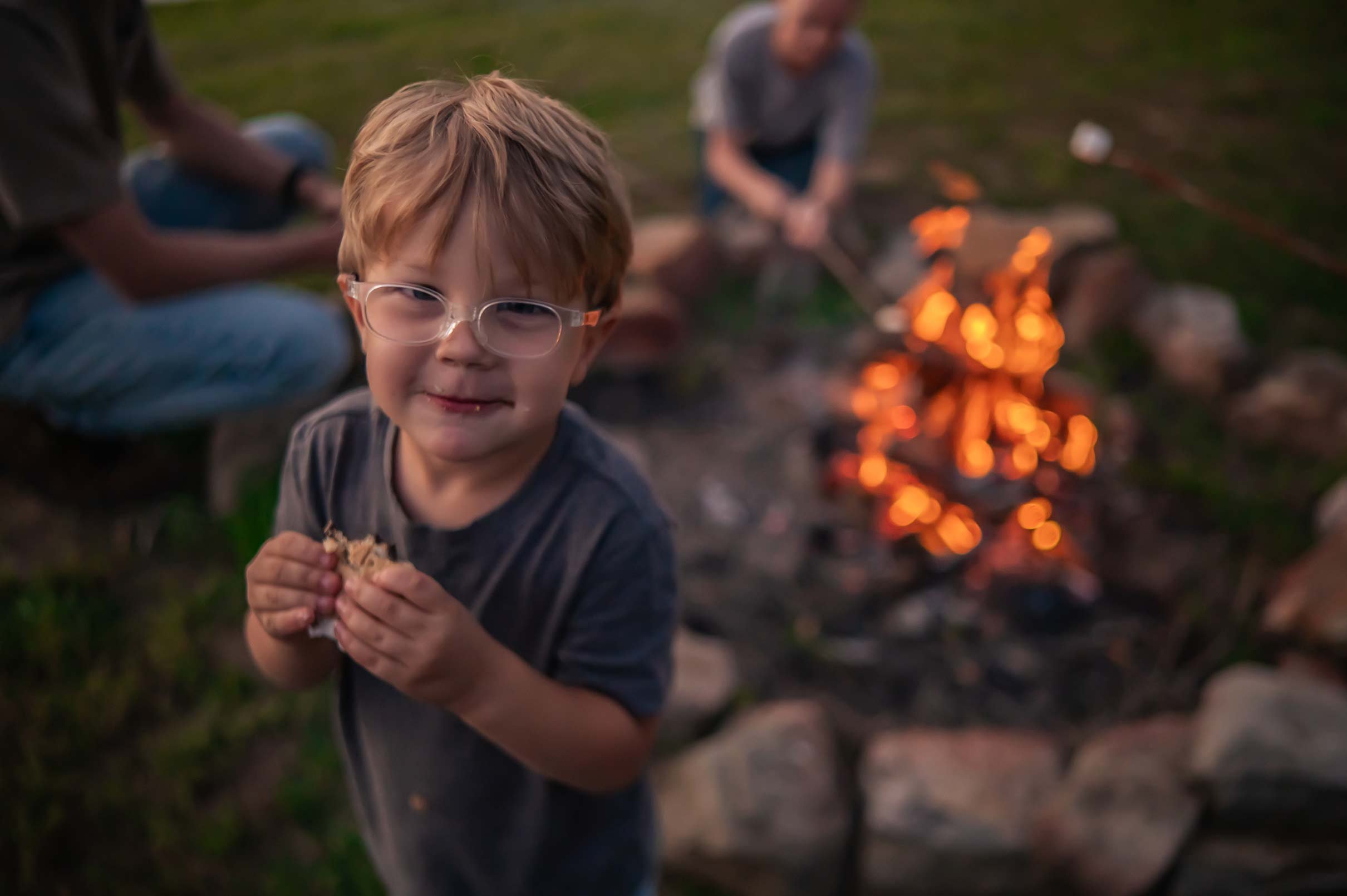 Young boy with glasses holding food, smiling at camera, outdoors during evening around a campfire.