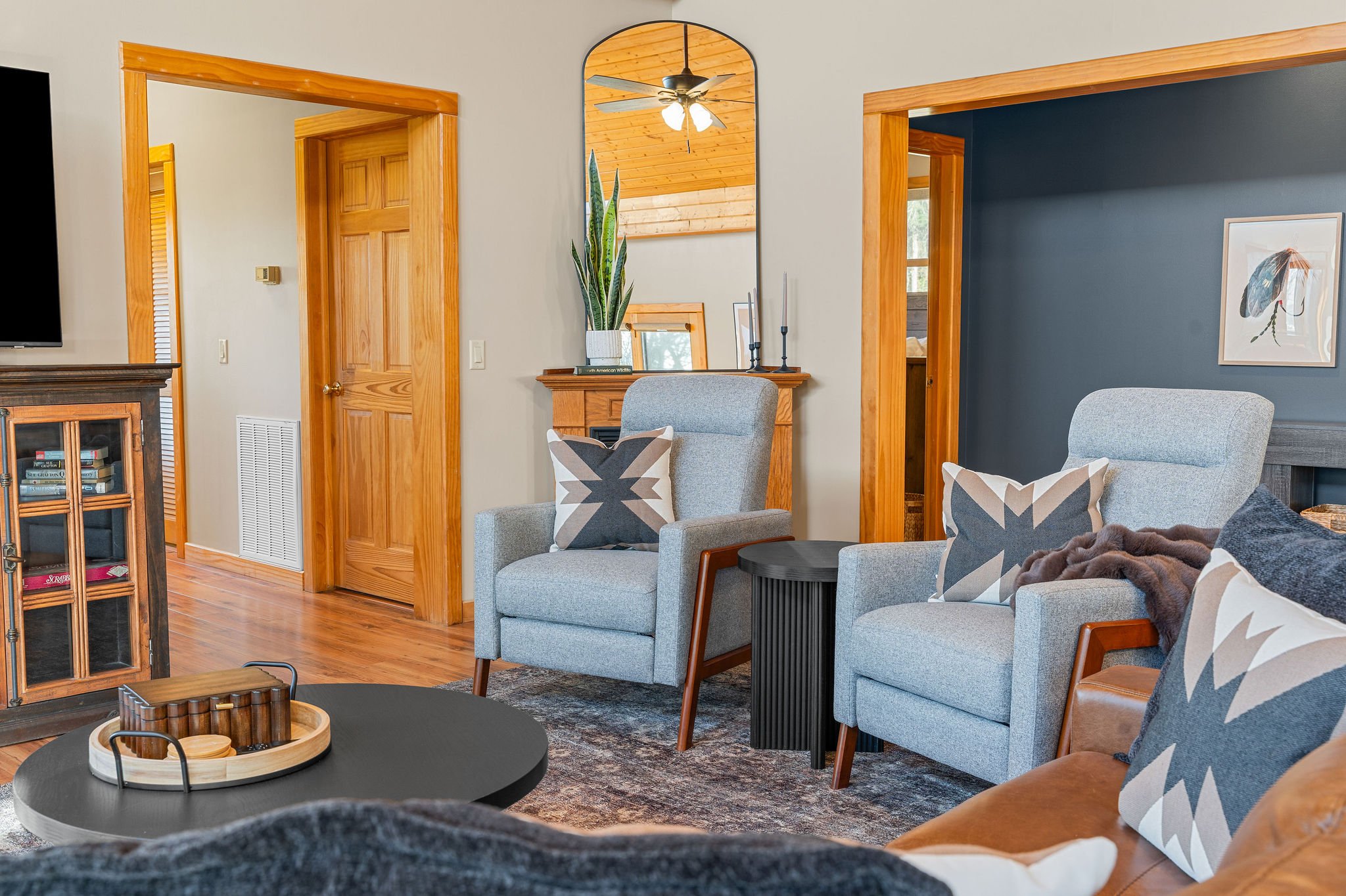Living room with light-colored walls, wooden door frames, a mirror, plants, armchairs with pillows, a round table, and a wall with blue and beige paint.