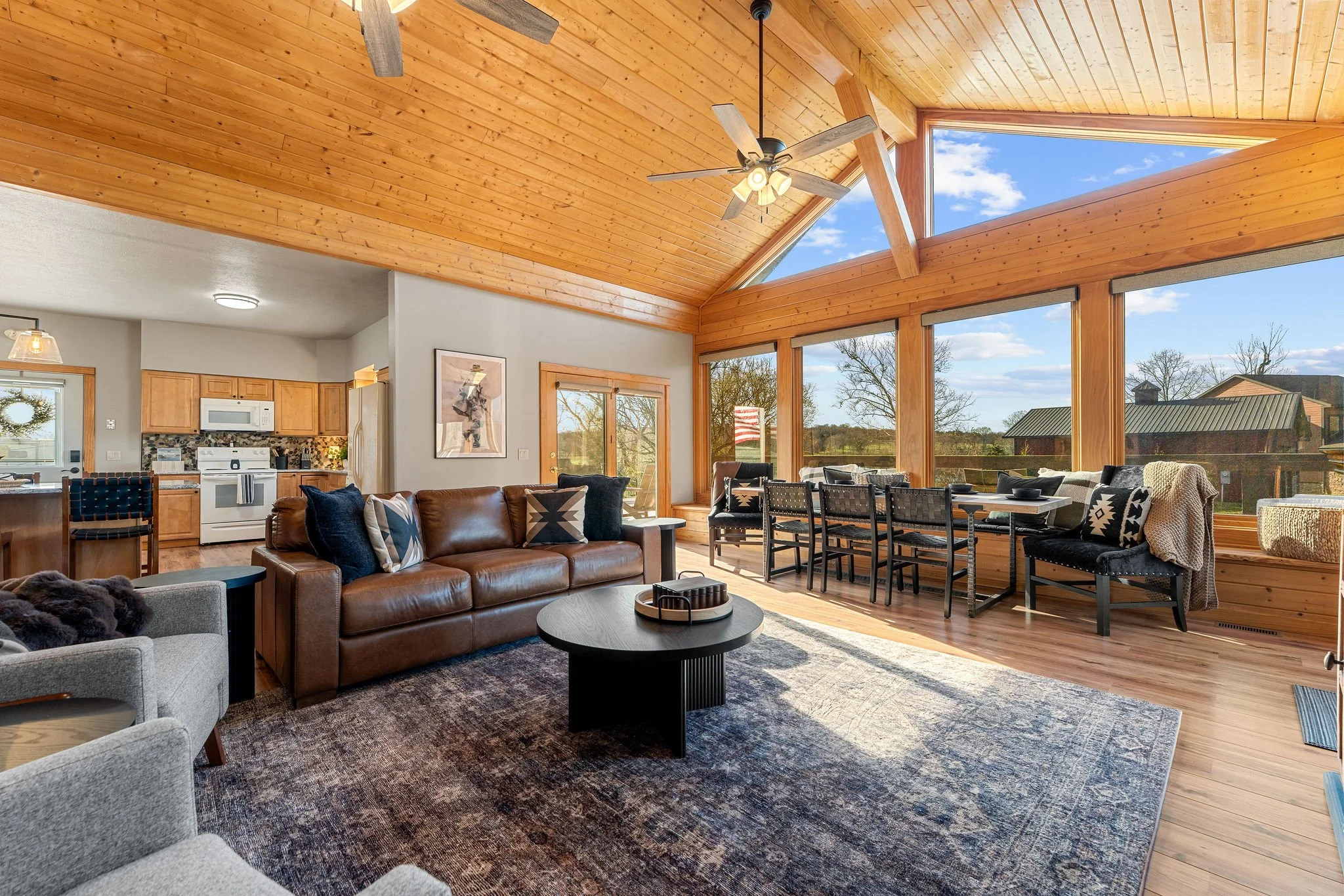 Living room with wood-paneled vaulted ceiling, ceiling fan, large windows showing a blue sky and trees, gray and brown furniture, and an open kitchen area.