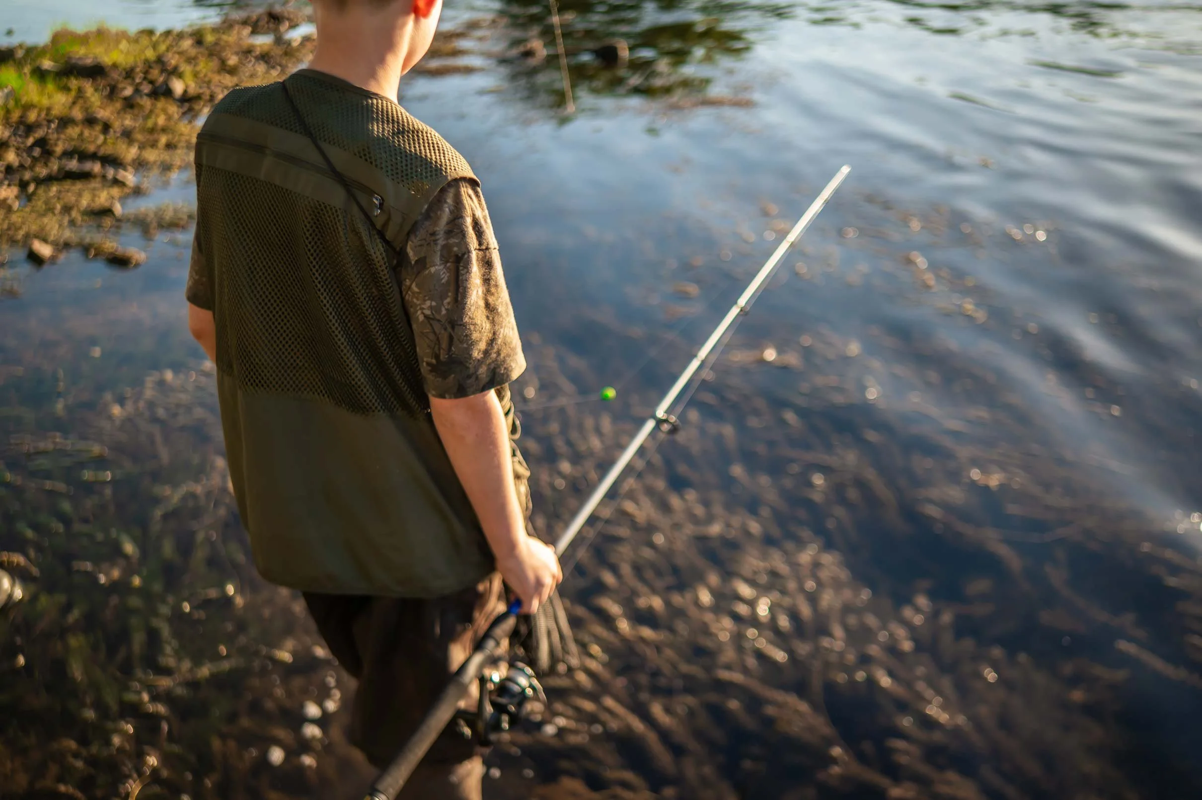 A young boy fishing at the edge of a body of water, standing on the shore during late afternoon, wearing a camouflage shirt and looking at the water.