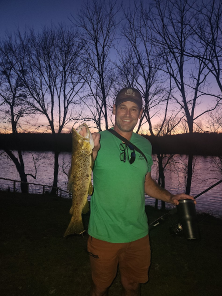 Man holding a large fish outdoors at sunset by a lake, with leafless trees in the background.