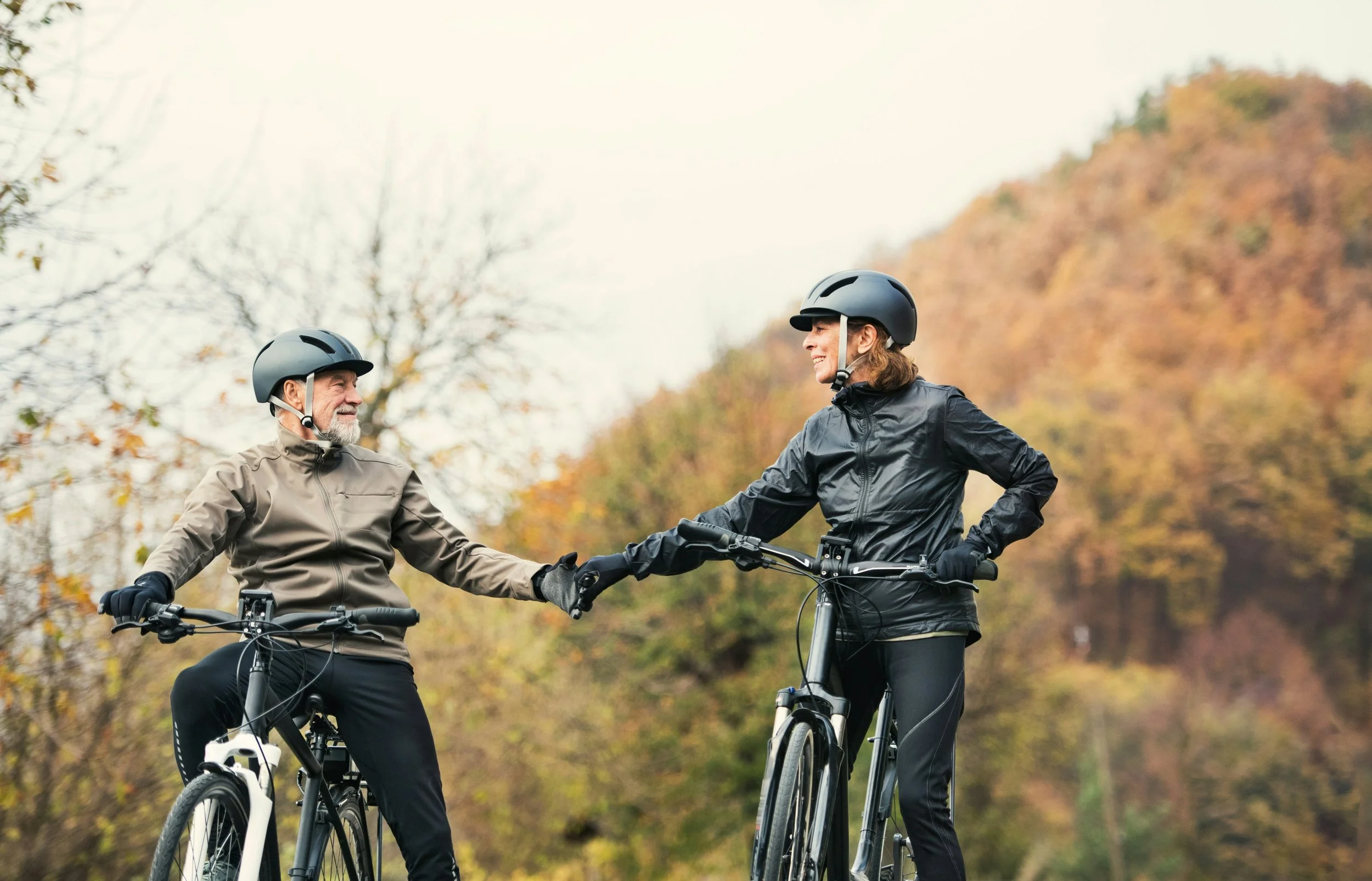 An older man and a younger woman riding mountain bikes outdoors, holding hands and smiling, with autumn trees in the background.