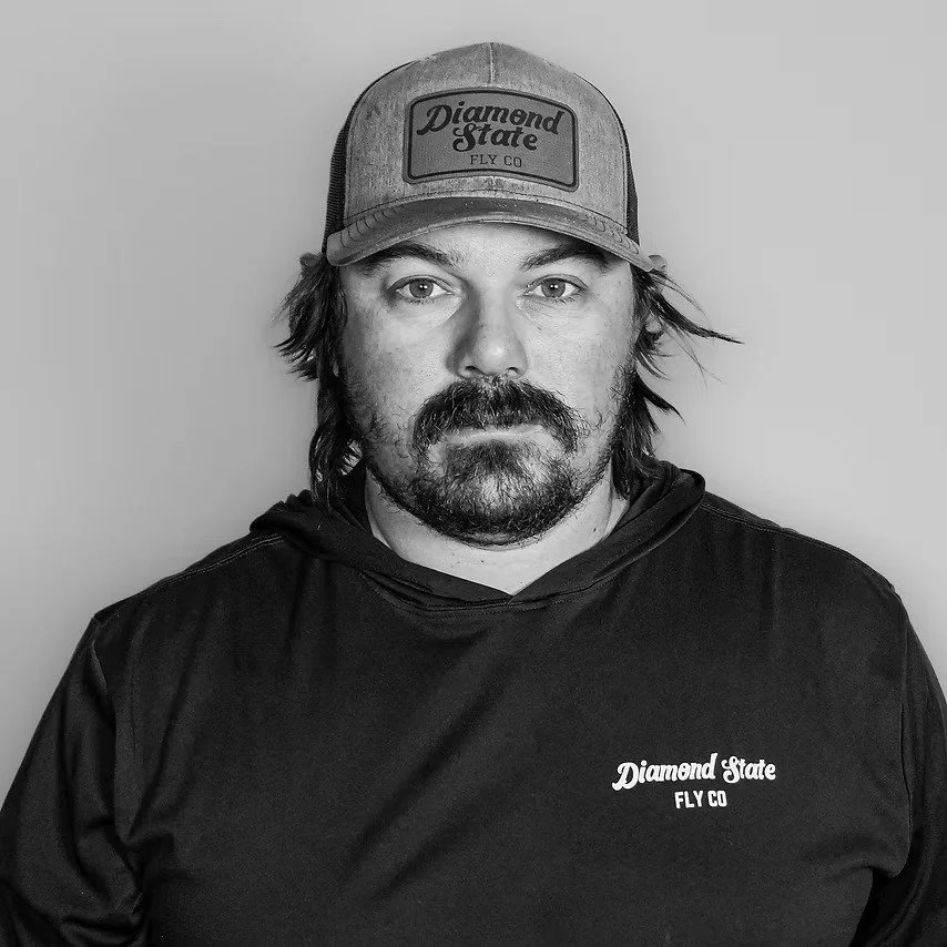 A man with a mustache and beard wearing a baseball cap and a black hoodie with "Diamond State FLY CO" printed on it, standing against a plain background.