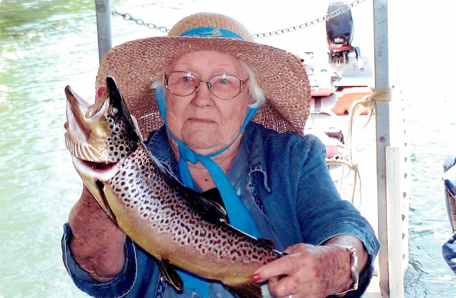 Elder woman wearing a wide-brimmed straw hat, glasses, and a denim shirt holding a large fish with spotted pattern while on a boat.