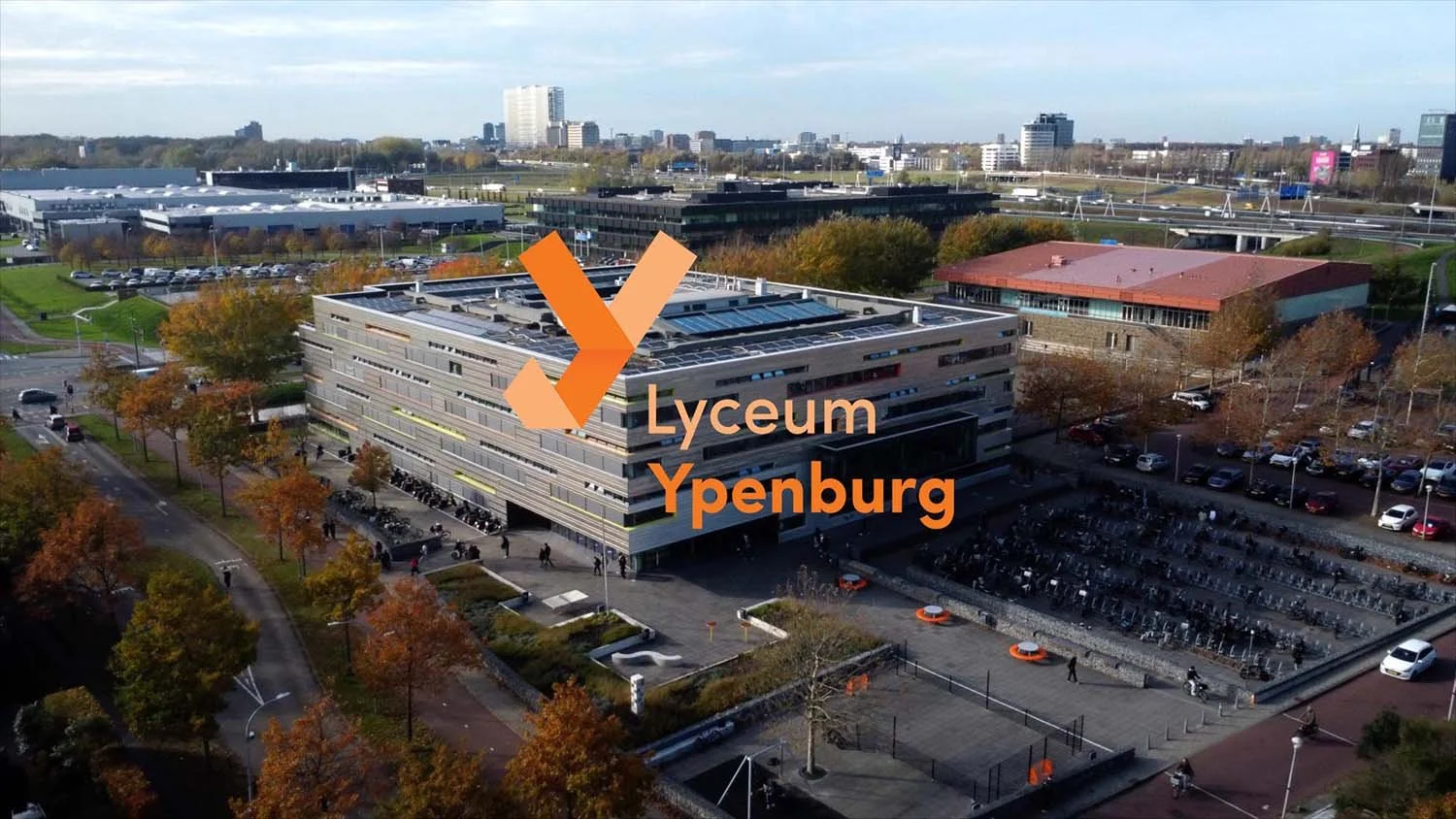 Aerial view of Lyceum Ypenburg building with surrounding trees in autumn, parking lot, and nearby streets in a city.