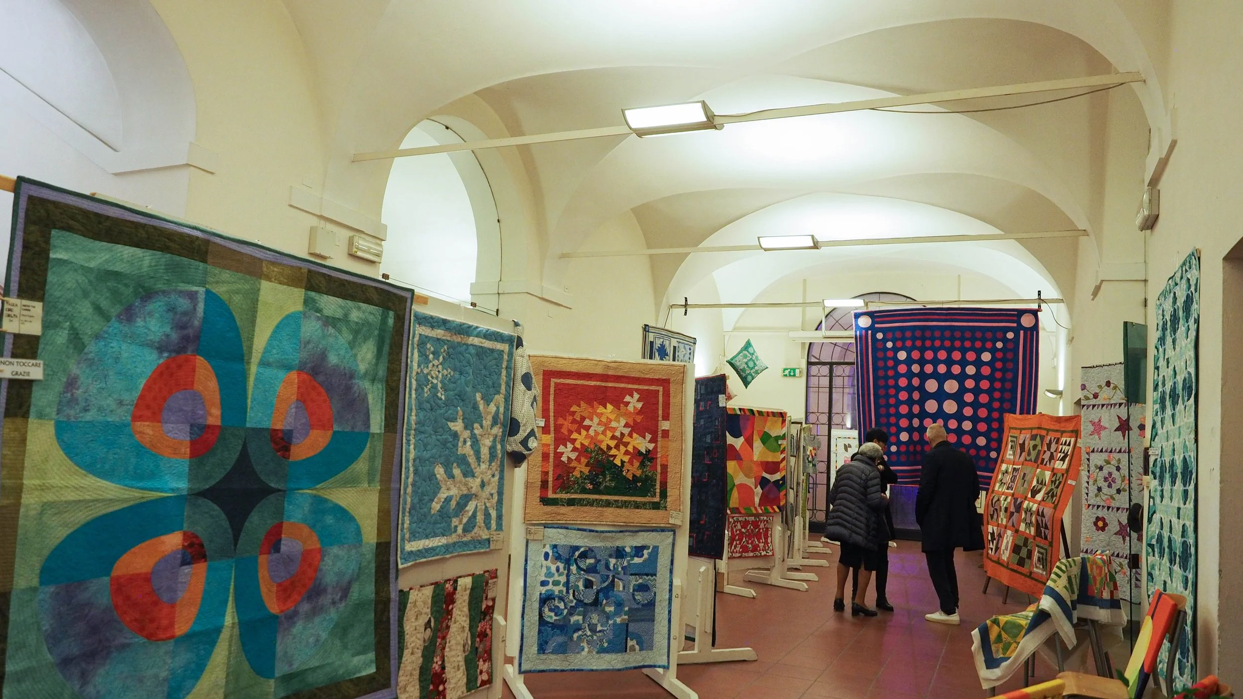 Mostra di trapunte colorate e artistiche esposte su supporti a forma di cavalletto, con persone che osservano e discutono delle opere in un ambiente di galleria o sala espositiva.