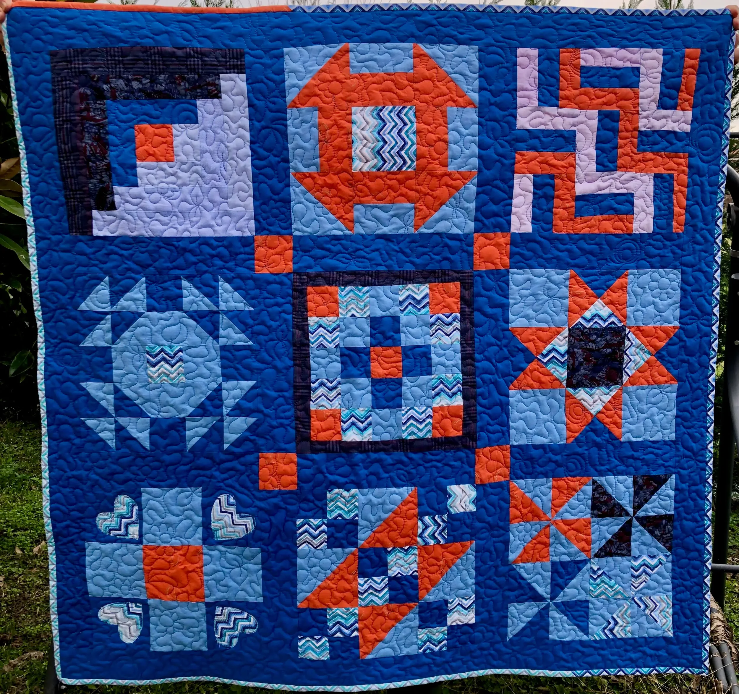 Copriletto patchwork con vari motivi geometrici in tonalità di blu, arancione, bianco e nero. Include stelle, cuori, quadrati, e forme astratte.