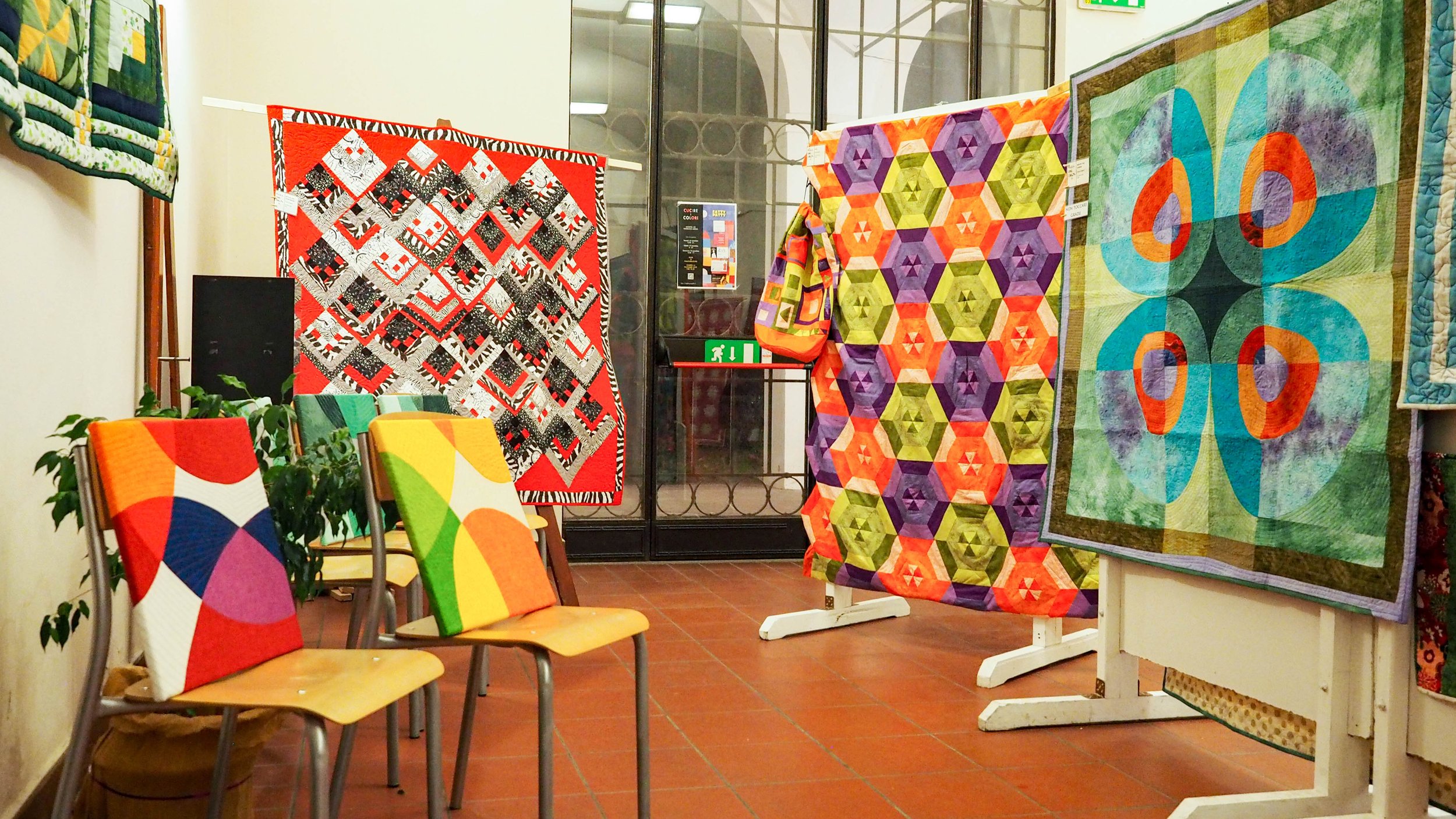 Mostra di tessuti e coperte colorate in un'esposizione, con sedie di legno e piante sullo sfondo.