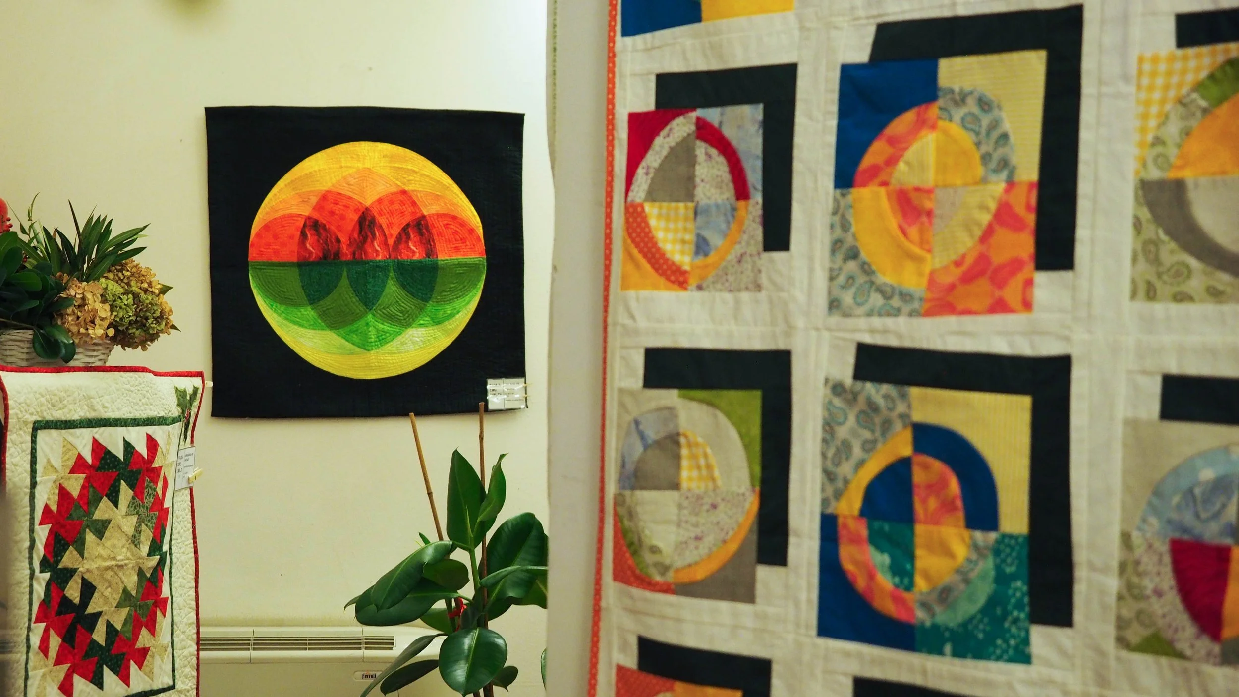 Arazzo composto da quadrati con disegni geometrici colorati e un quadro con un disegno circolare astratto con colori vivaci come giallo, arancione, verde, rosso, e blu, in un'area di esposizione.