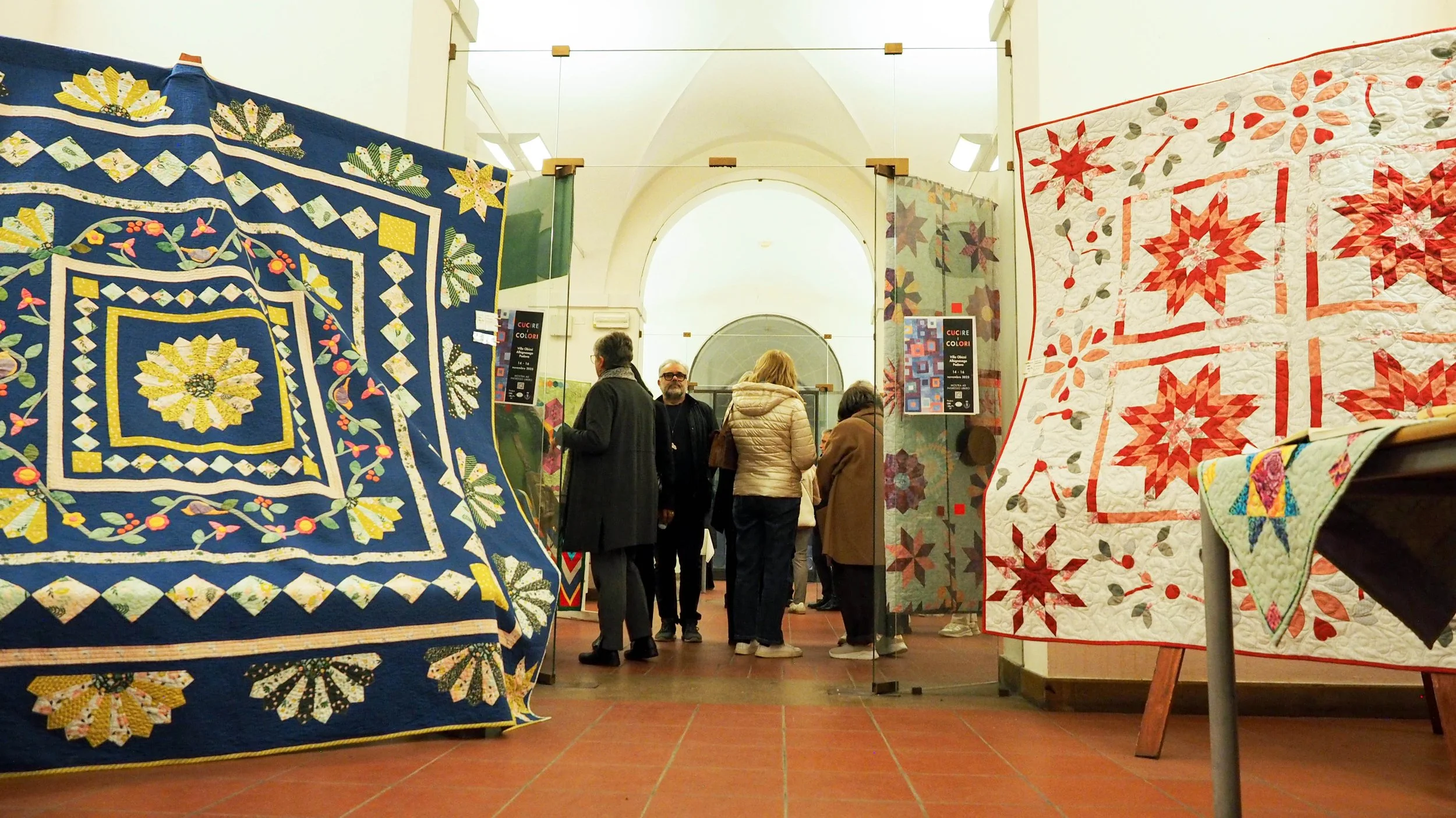 Mostra di coperte e quilt ricamati e lavorati a mano in un museo, con persone che osservano e discutono.