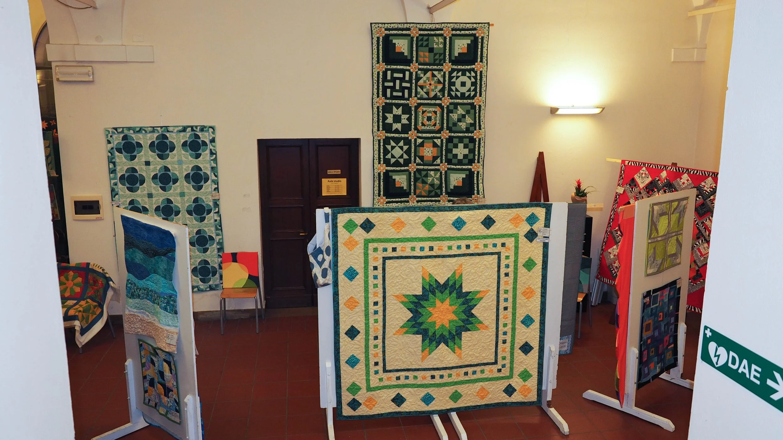 Mostra di diversi quilt colorati e decorativi in una sala espositiva con pareti bianche e illuminazione soffusa.