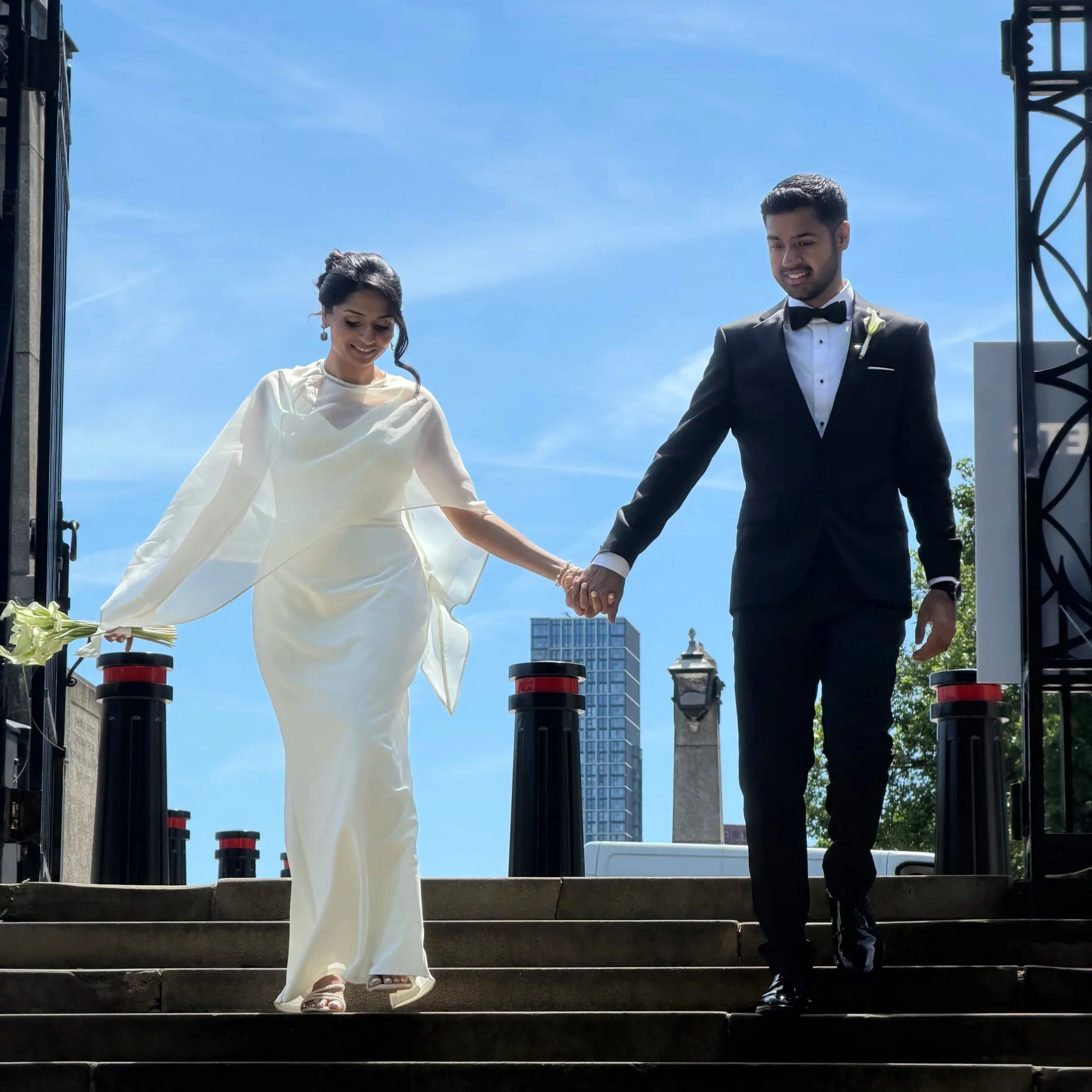 A bride and groom holding hands and walking down steps outdoors, with a blue sky and city buildings in the background, during a wedding ceremony.