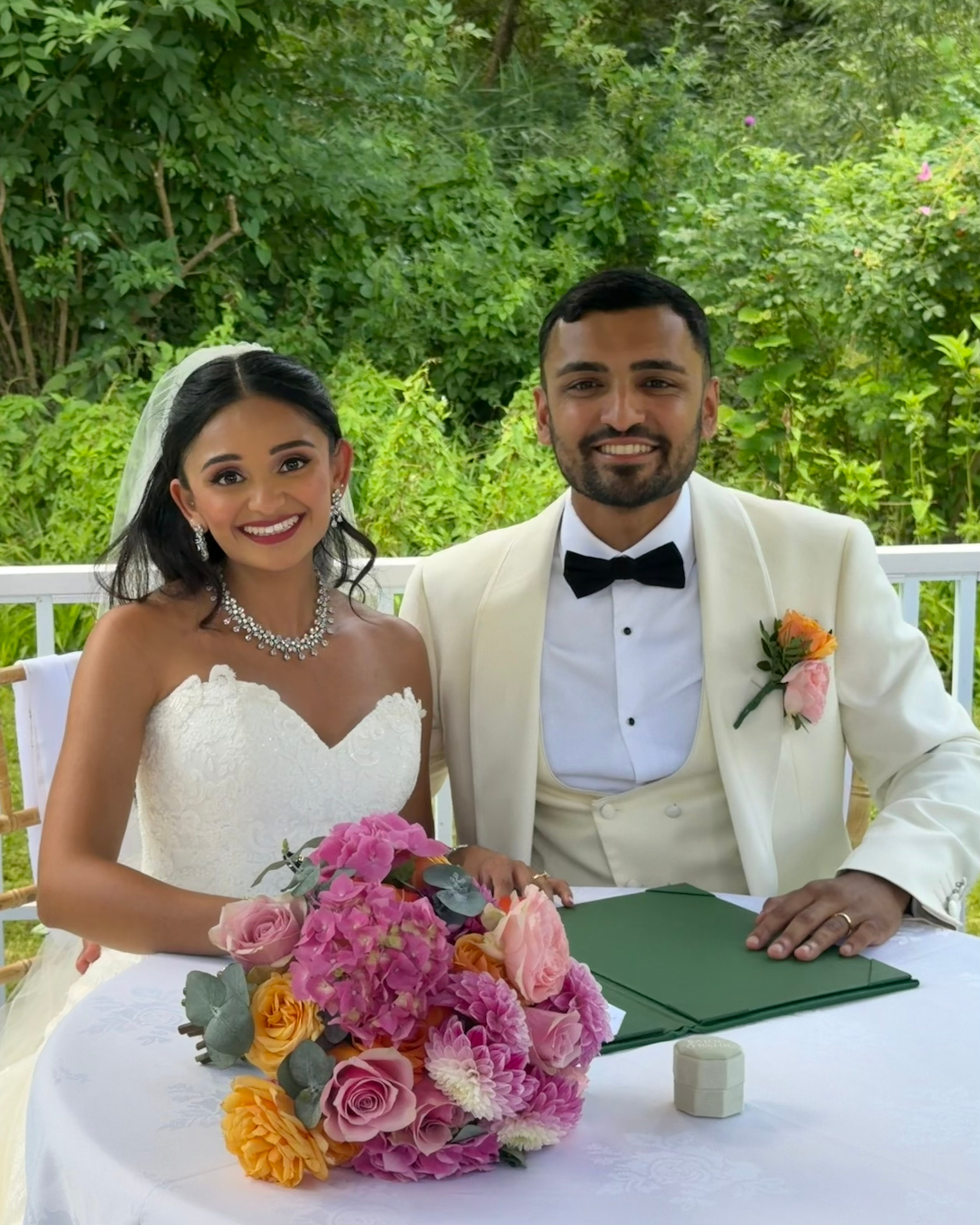 A bride and groom sitting at a wedding table, smiling, with a bouquet of pink, yellow, and peach flowers in front of them, against a green outdoor background.