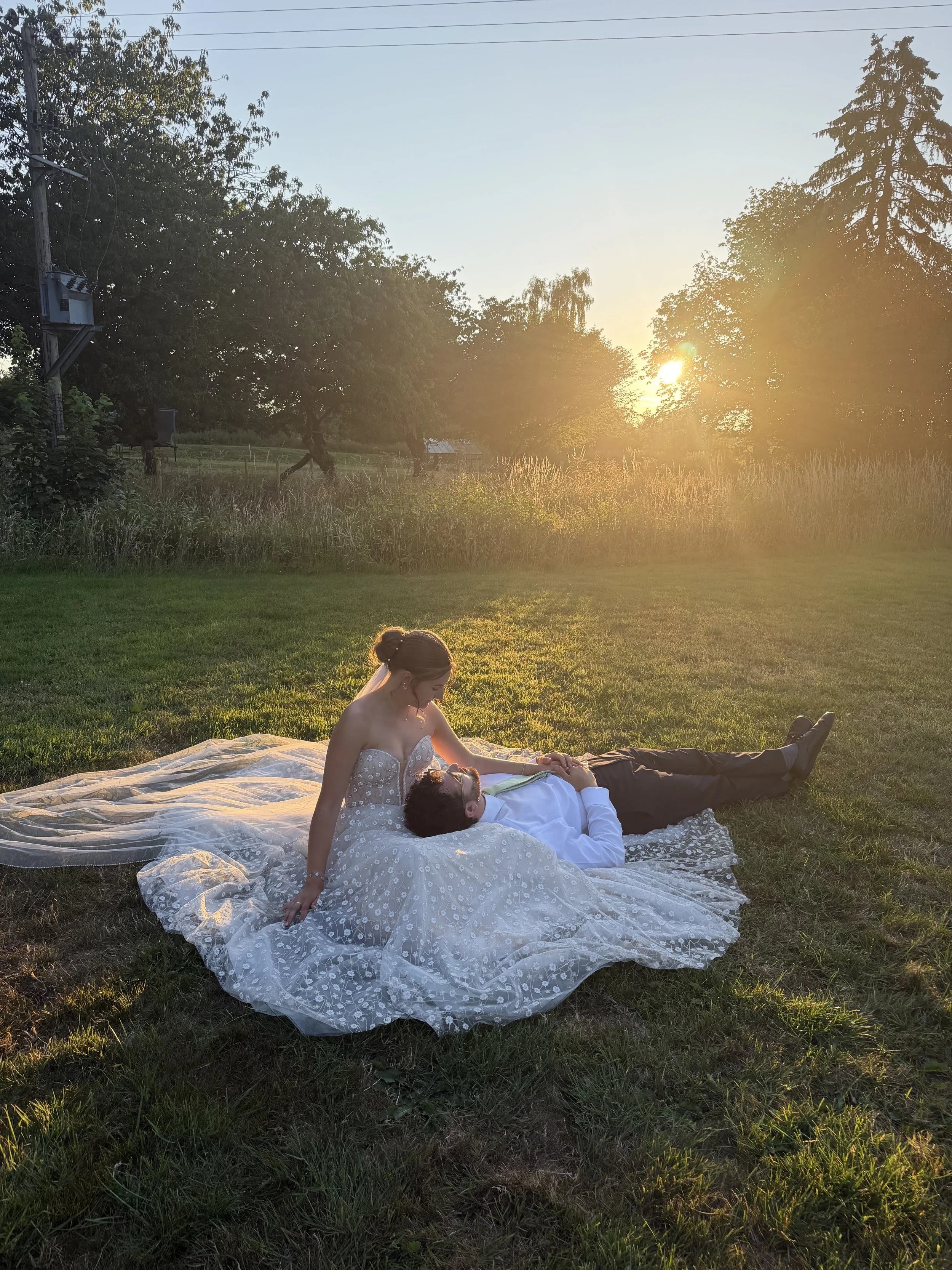 A couple lying on a blanket in a grassy field during sunset, surrounded by trees.