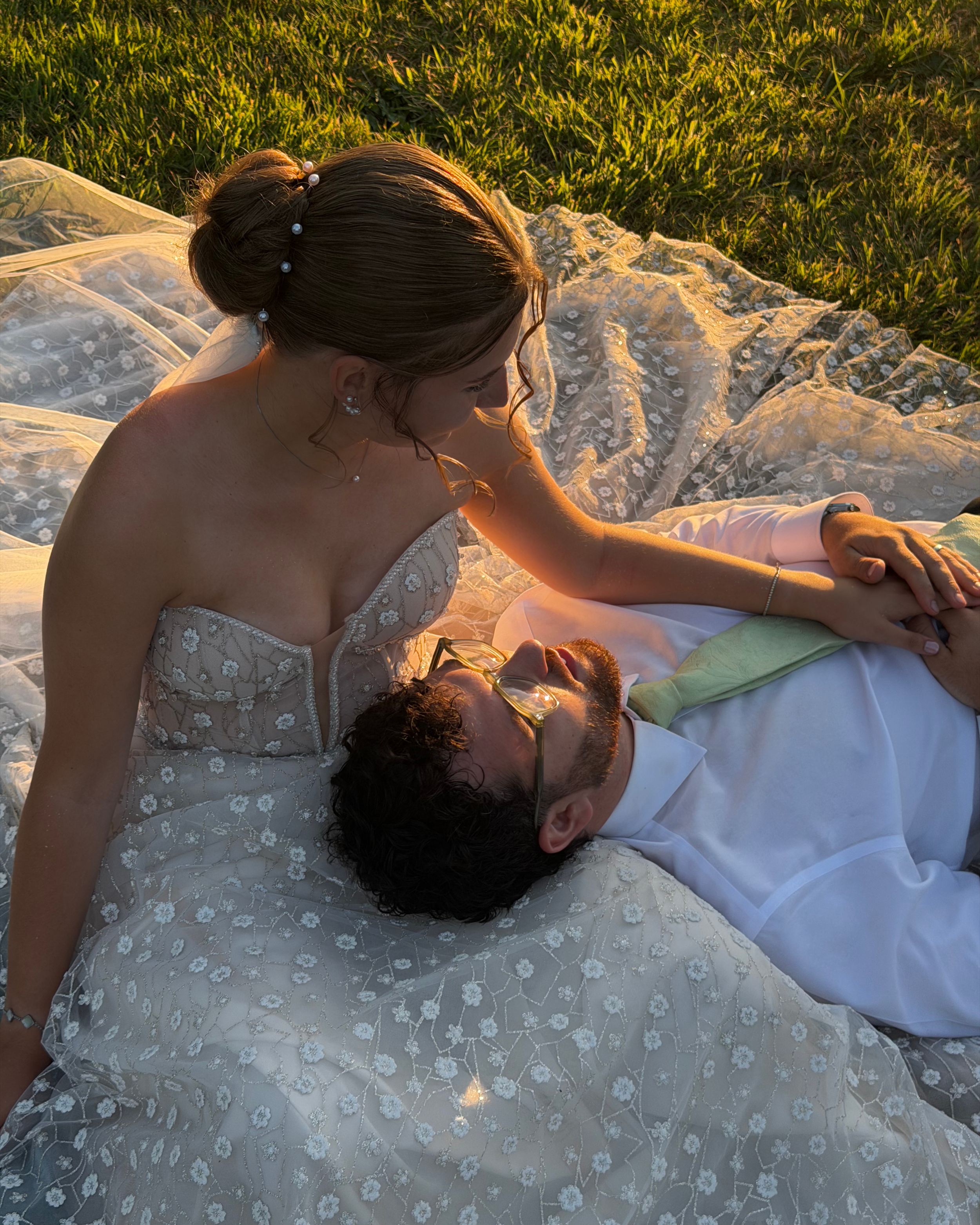 A bride and groom lying on a lace-covered surface outdoors during sunset, with the bride sitting up and the groom lying down beneath her, sharing an intimate moment.