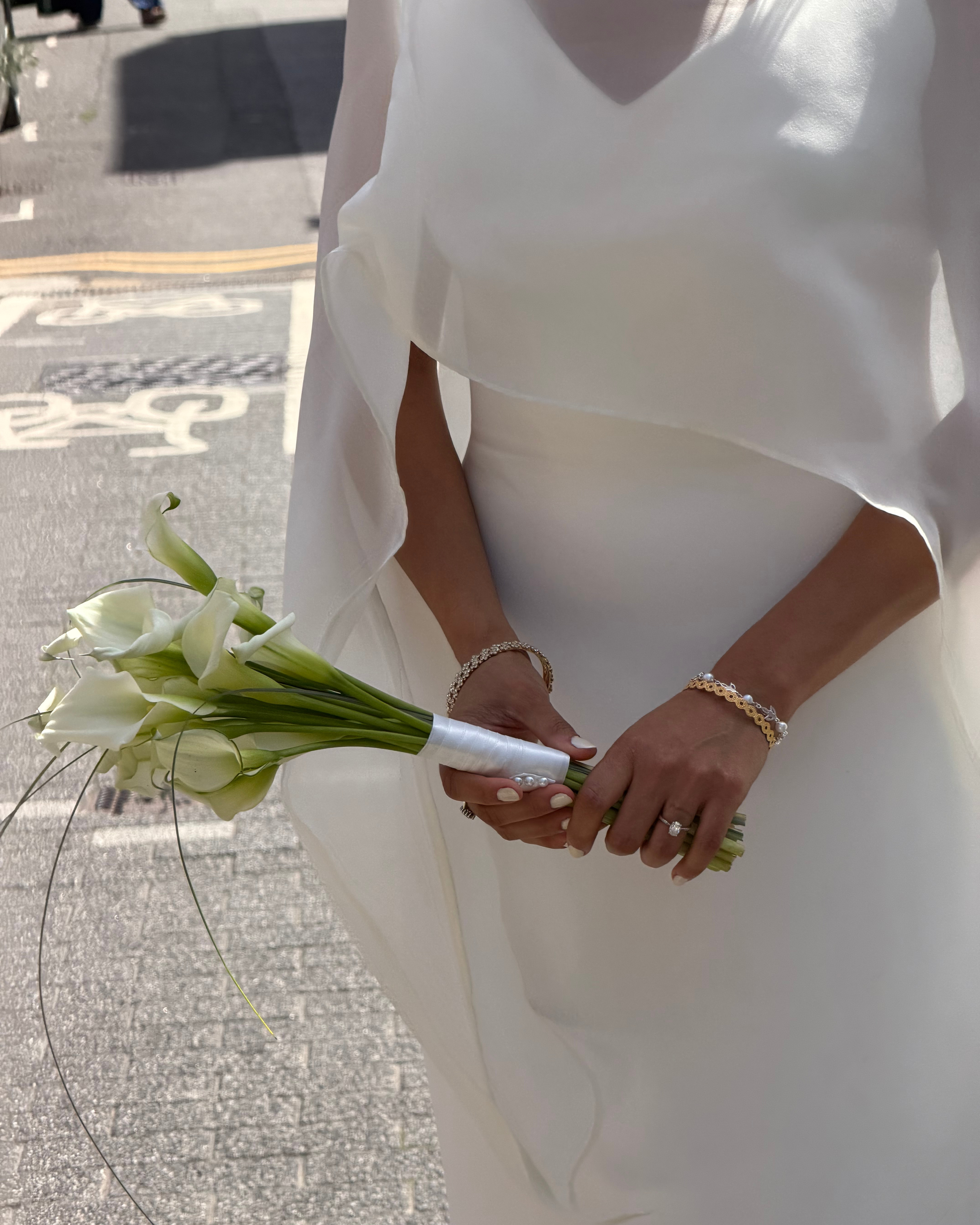 A bride wearing a white wedding dress holding a bouquet of white calla lilies, with her hands adorned with jewelry, including a ring and bracelets, standing outside near a sidewalk.