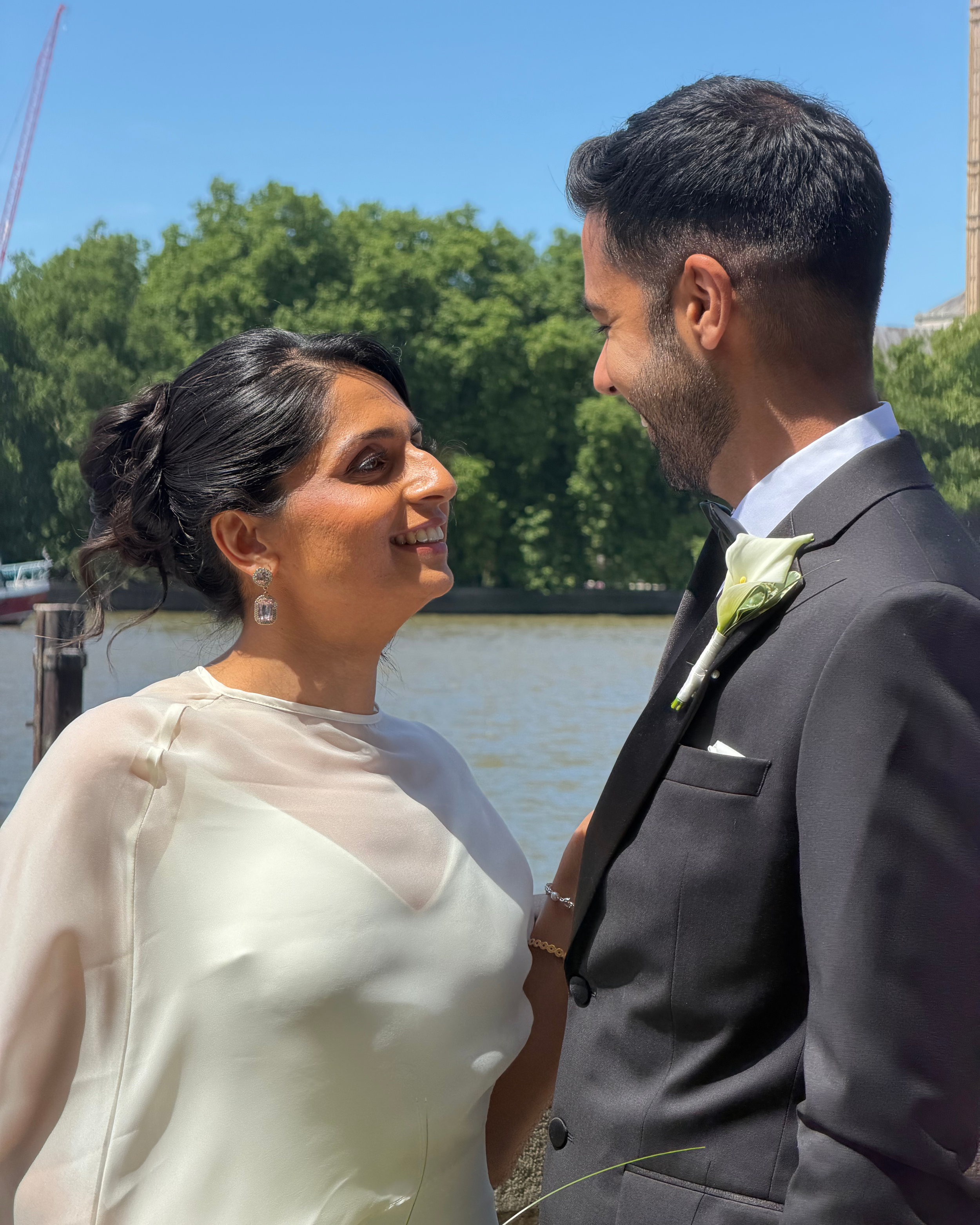 A bride and groom facing each other, smiling, standing outdoors near water with trees in the background, during their wedding ceremony.