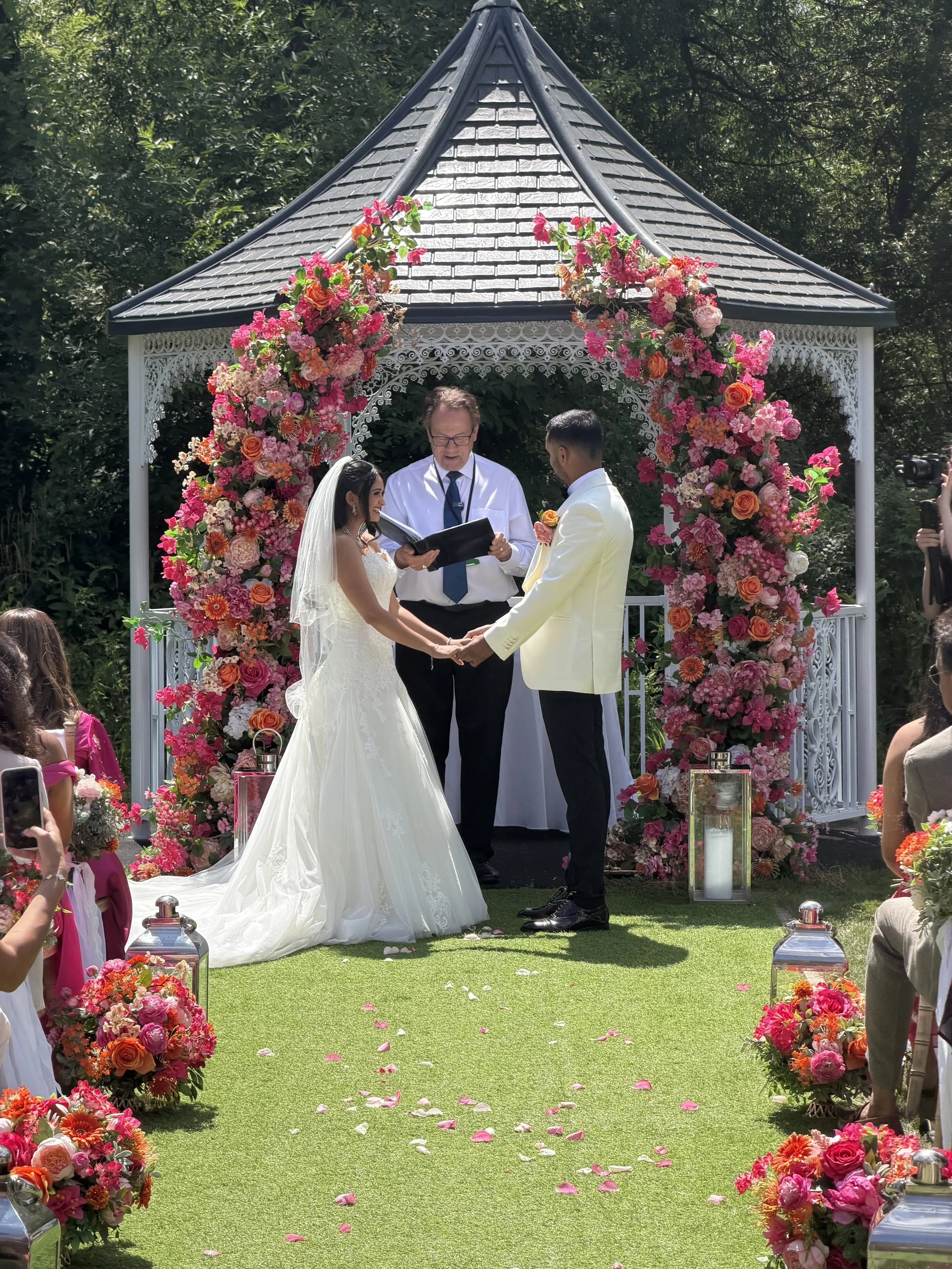 A wedding ceremony taking place outdoors under a decorated gazebo with pink and orange flowers. The bride and groom are holding hands, facing each other, with a officiant reading from a book behind them. Guests are seated around, and flower arrangements line the aisle.