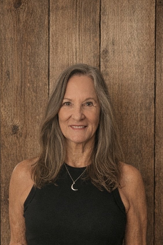 A smiling  woman with long hair, wearing a black sleeveless top and a moon-shaped necklace, standing in front of a wooden wall background.