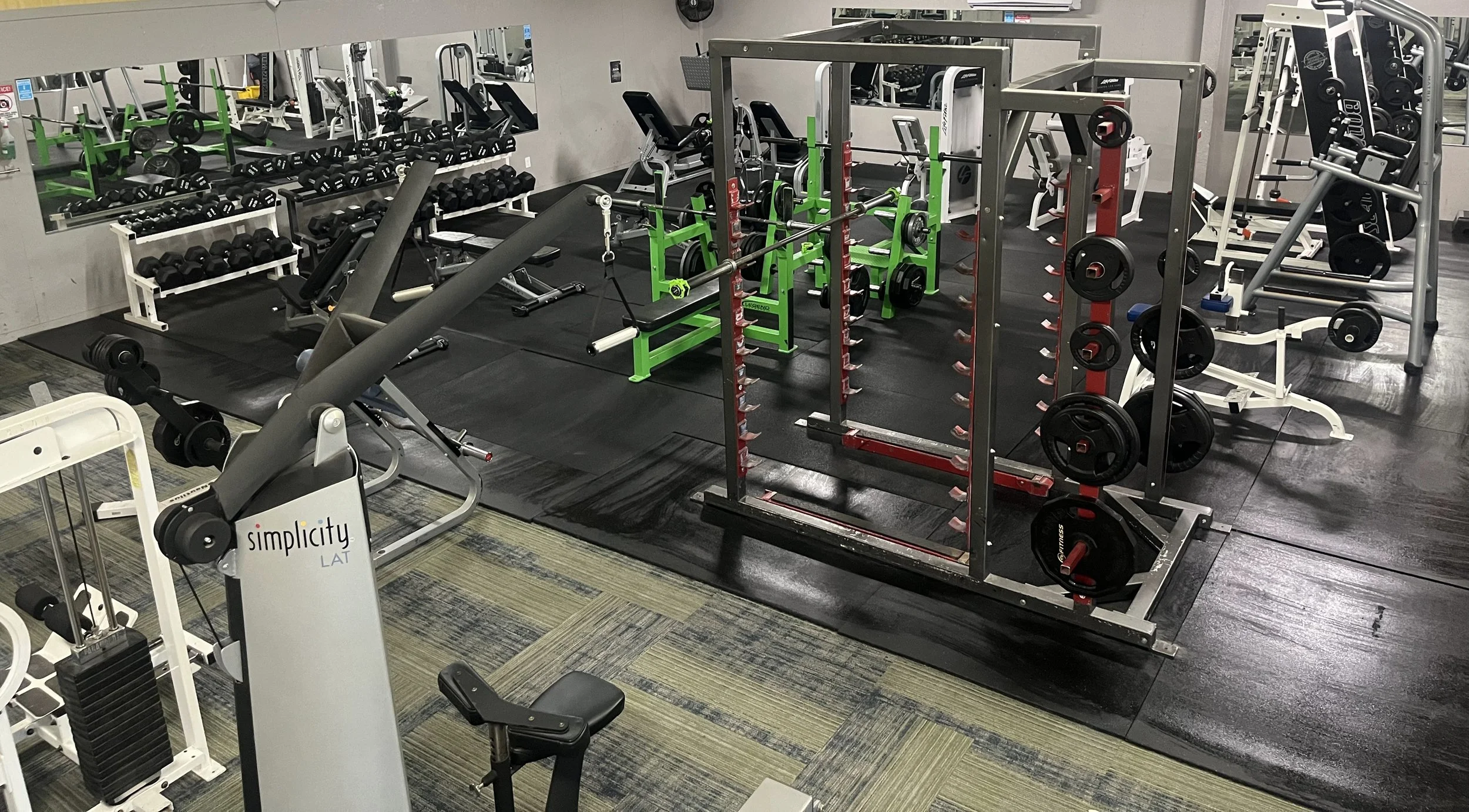 Interior view of a gym with various weight lifting equipment and exercise machines, including dumbbells, barbells, squat racks, and benches.