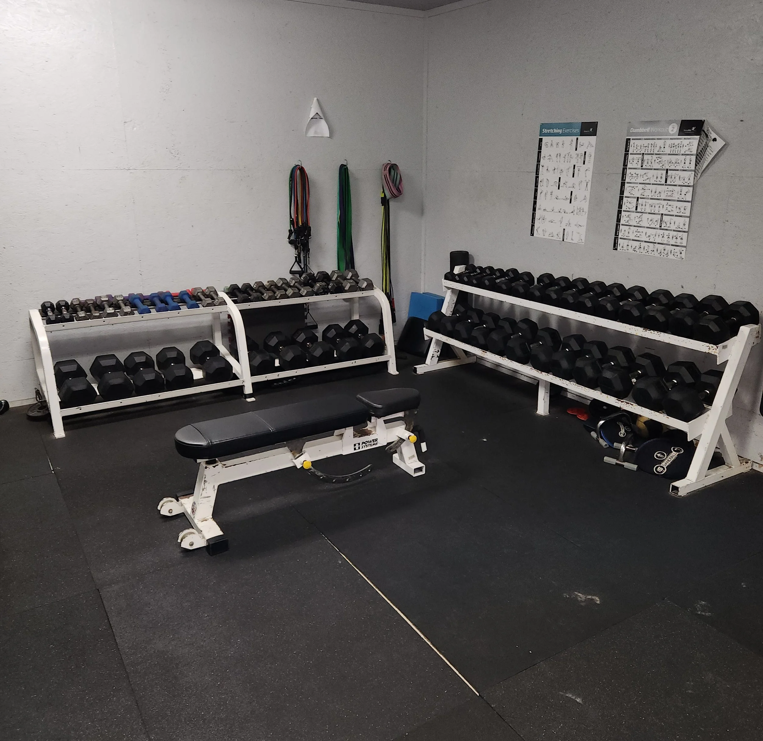A small gym corner with two white racks of black dumbbells, a black adjustable workout bench, resistance bands hanging on the wall, and exercise posters.