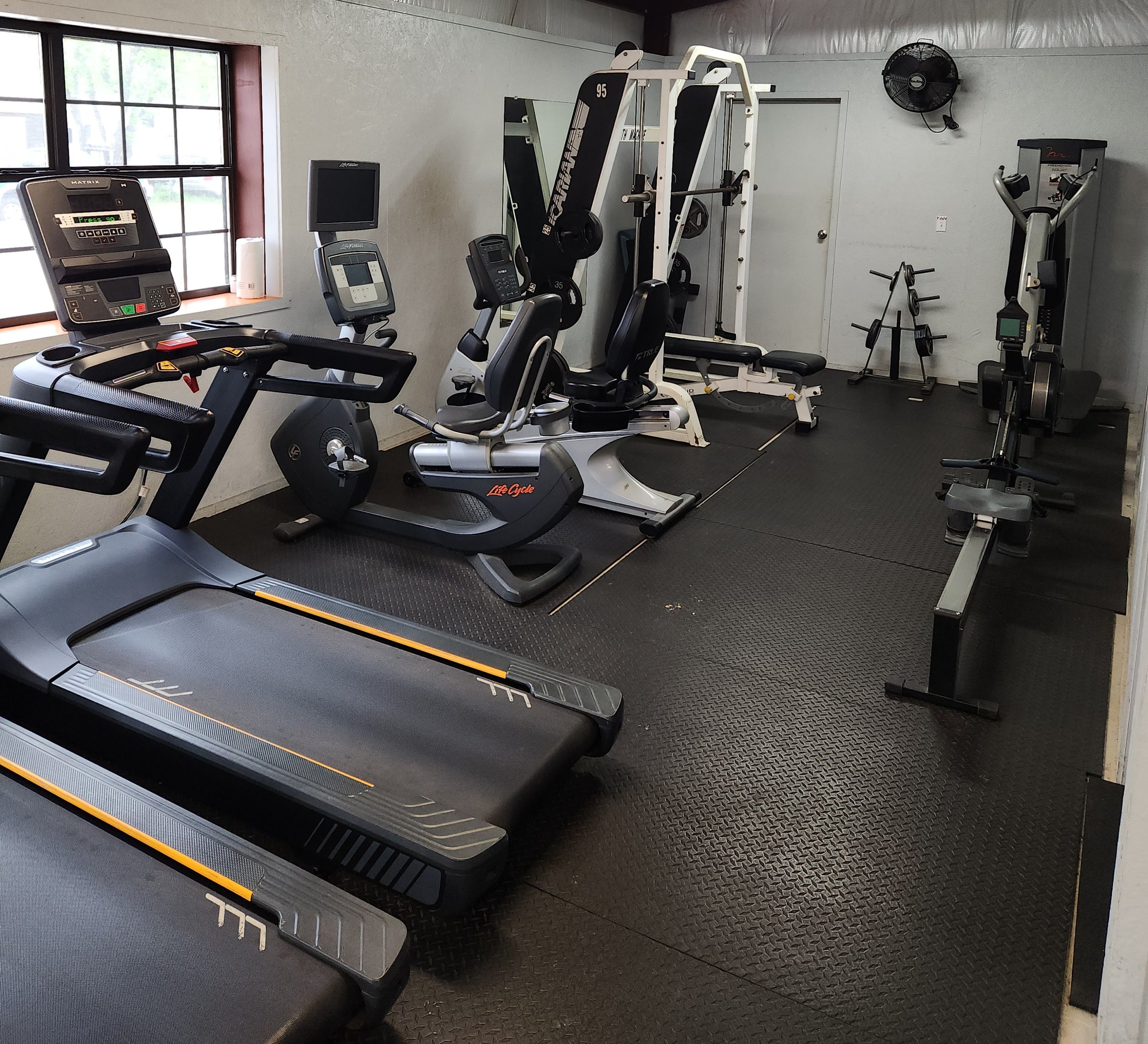A small gym with exercise equipment including treadmills, stationary bikes, an elliptical, and weight machines, with black rubber flooring and a window.