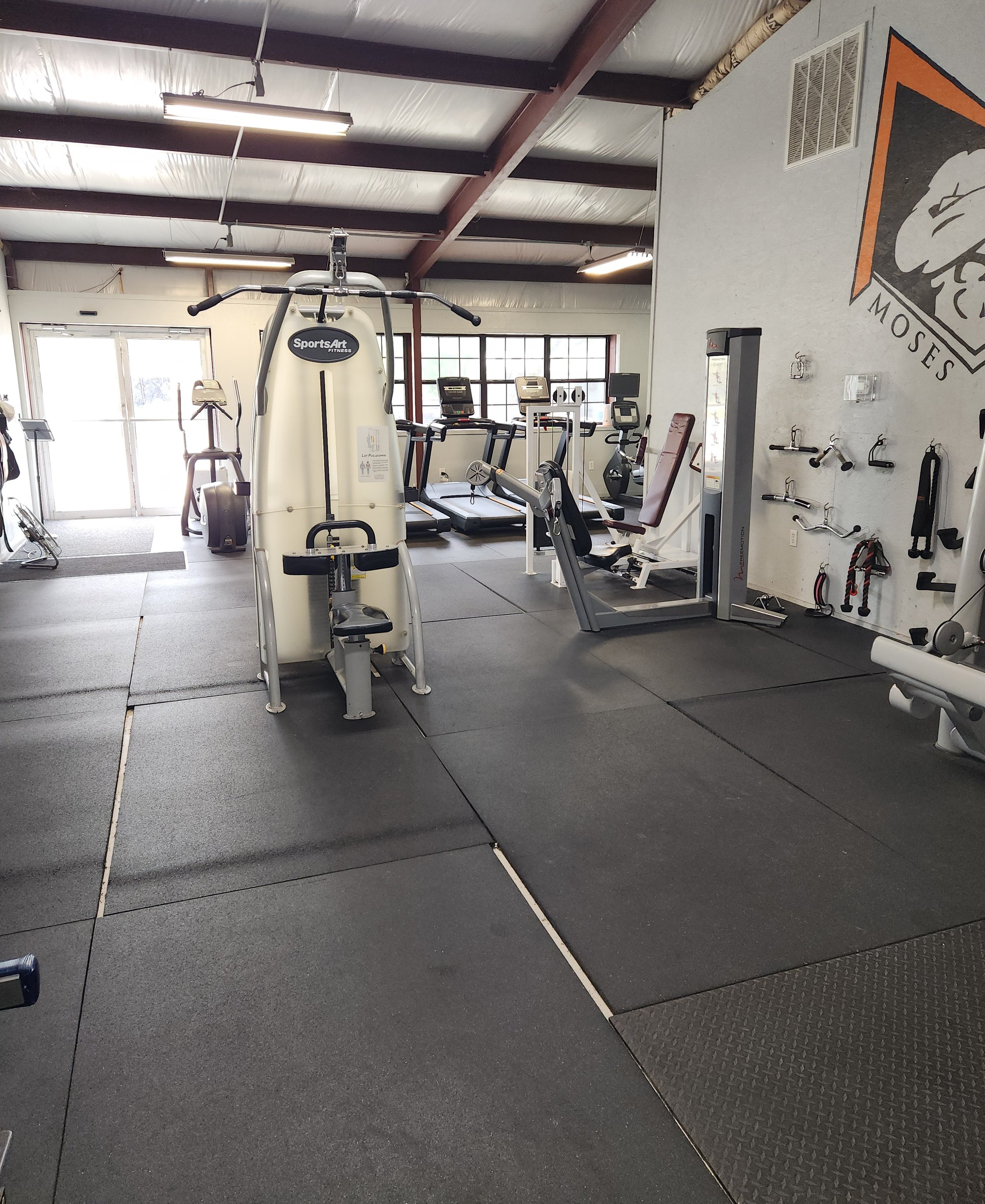 Empty gym with workout equipment including a seated rowing machine, treadmills, and various fitness tools against a wall with a Moses logo.
