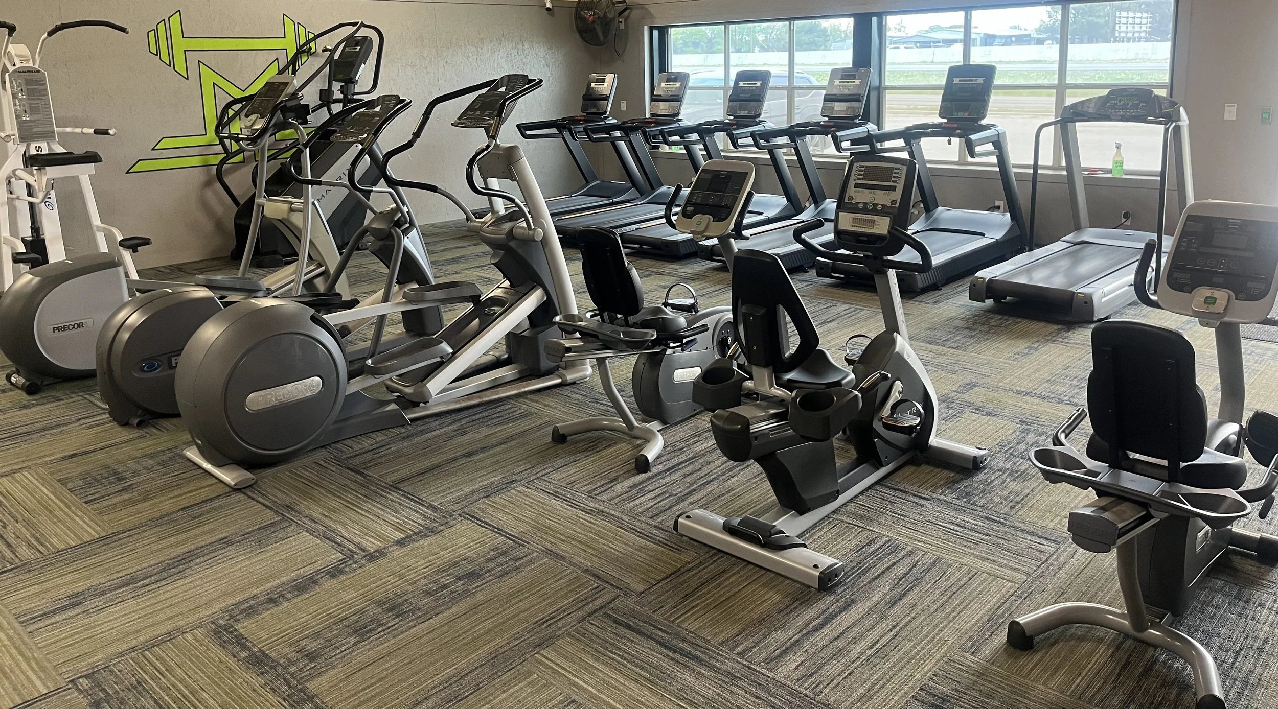 Indoor gym with various cardio equipment including ellipticals, treadmills, and stationary bikes, in front of large windows.
