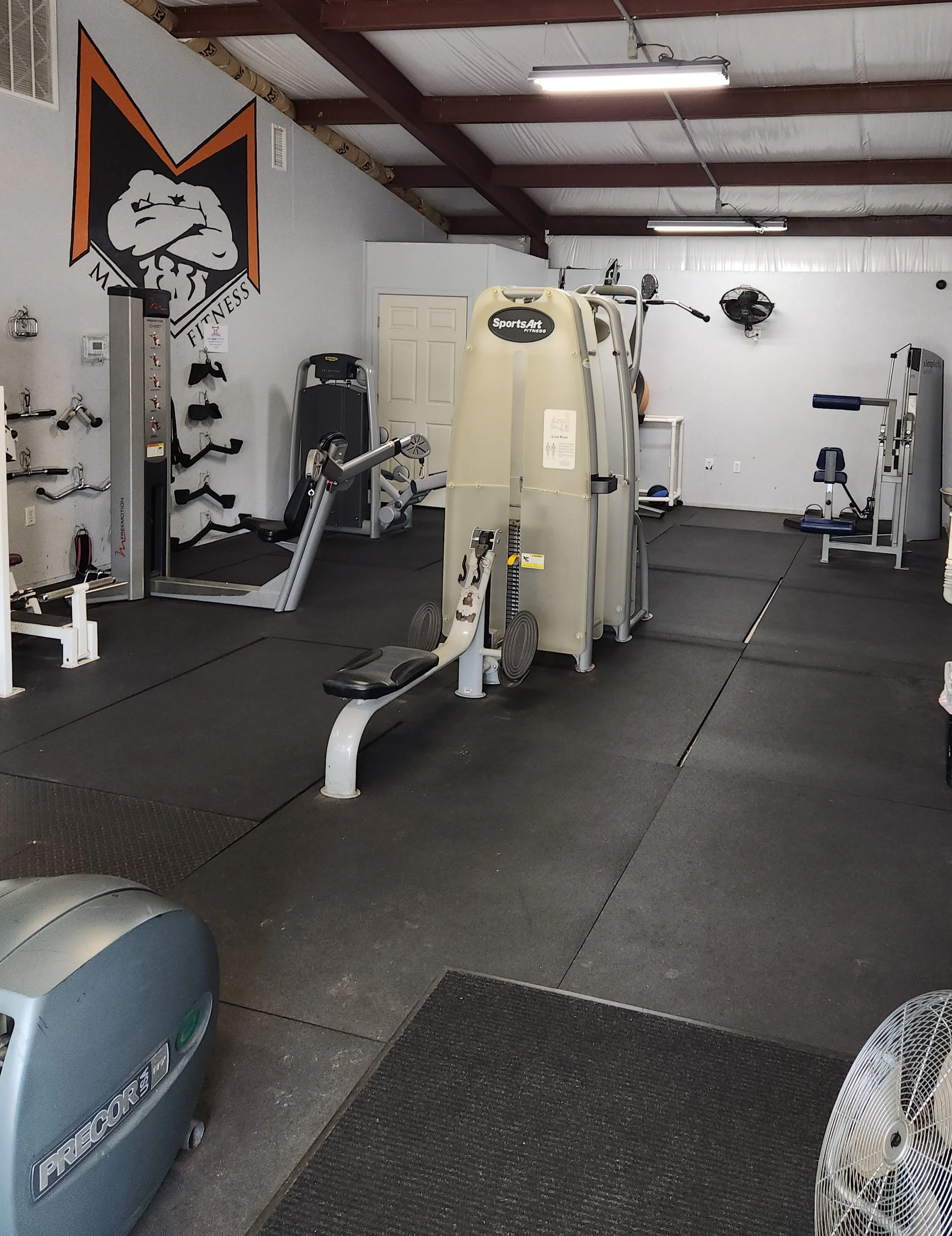 Interior view of a gym with various exercise equipment, including a shoulder press machine, a cardio machine, and weight racks, with a large logo on the wall depicting a clenched fist and the words "Momentum Fitness".