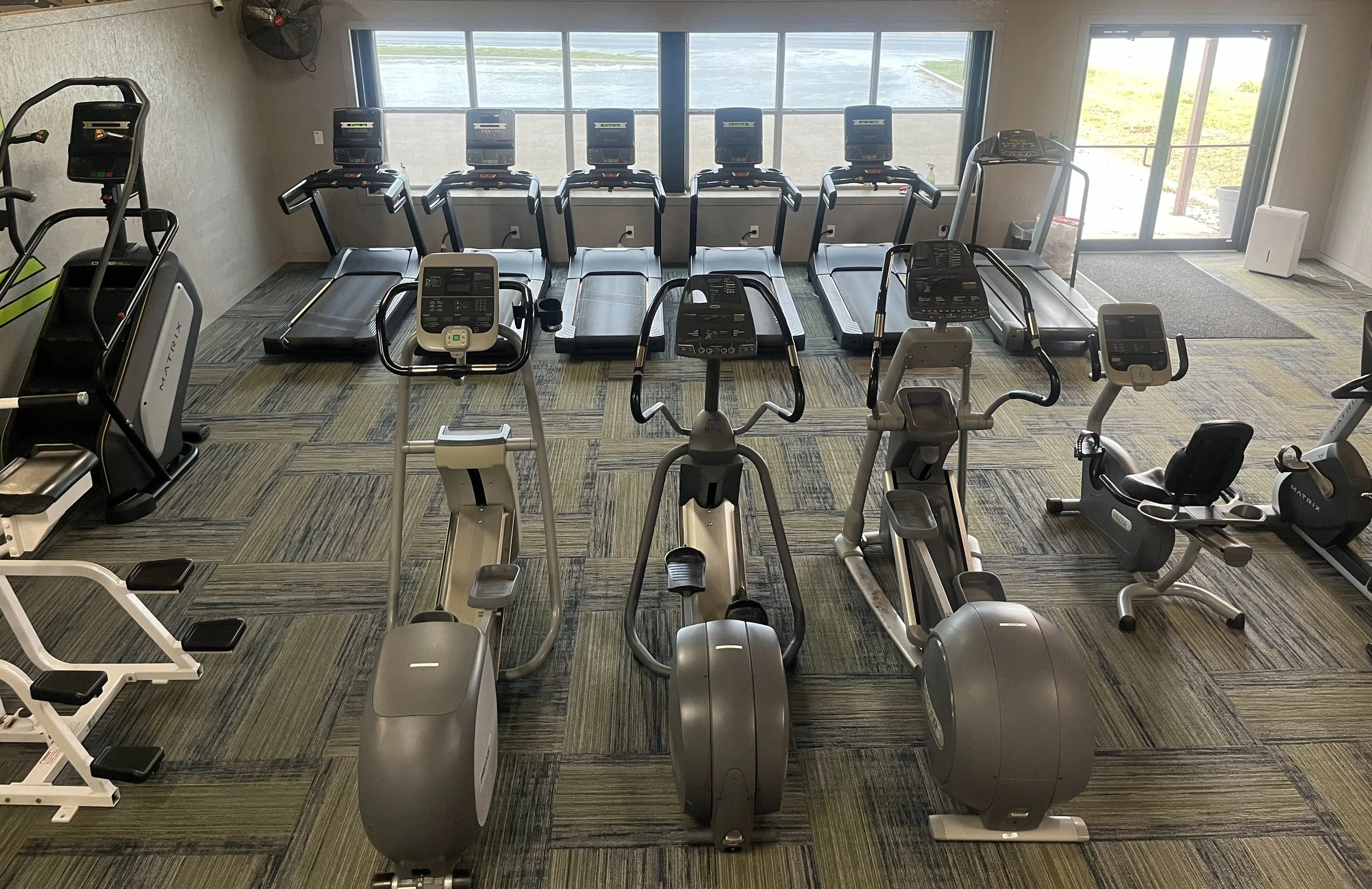 Indoor gym room with multiple treadmills, stationary bikes, elliptical trainers, and weight machines, large windows with an outdoor view.