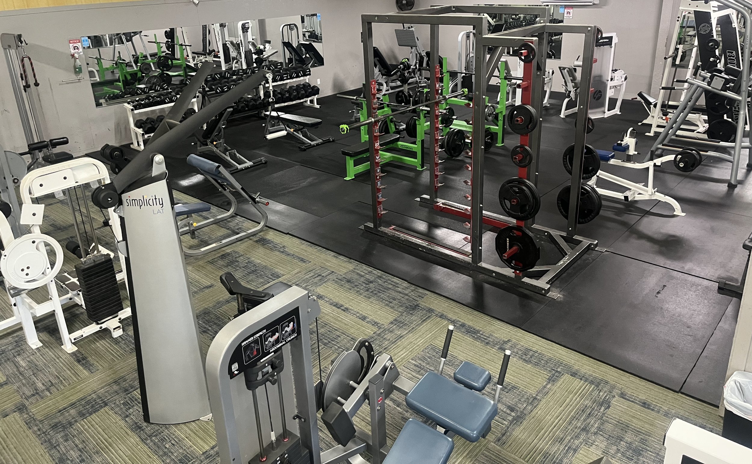 Gym with various workout equipment including weight machines, free weights, and benches, with a mirrored wall in the background.