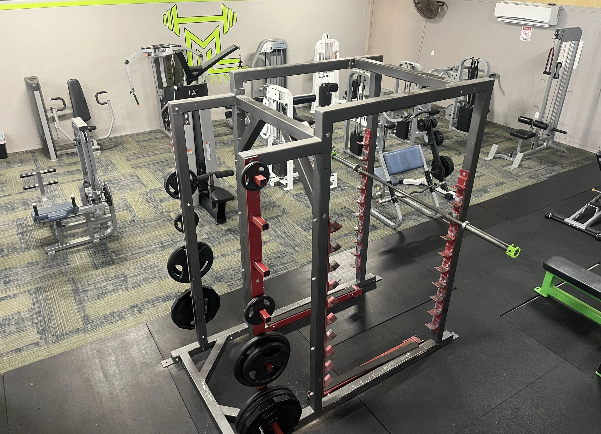 Indoor gym with various workout equipment including weight lifting stations, benches, and machines. The room has a black mat area and a carpeted section with a fitness logo on the wall.
