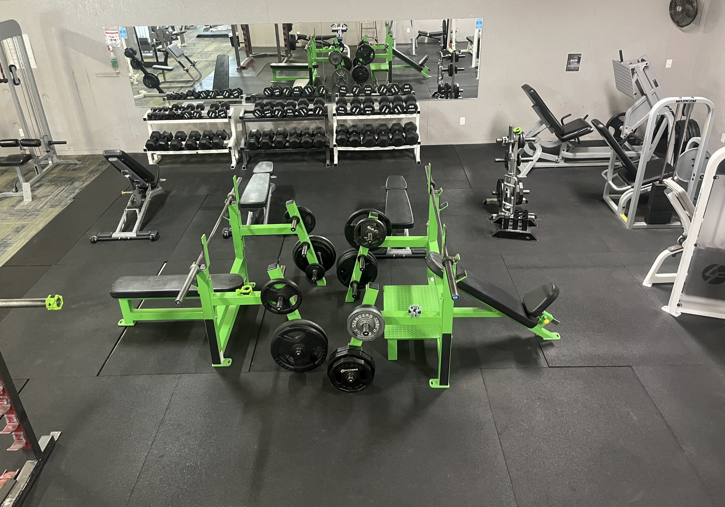 A gym with various workout equipment, including green benches, dumbbells, barbells, weight plates, and cardio machines.