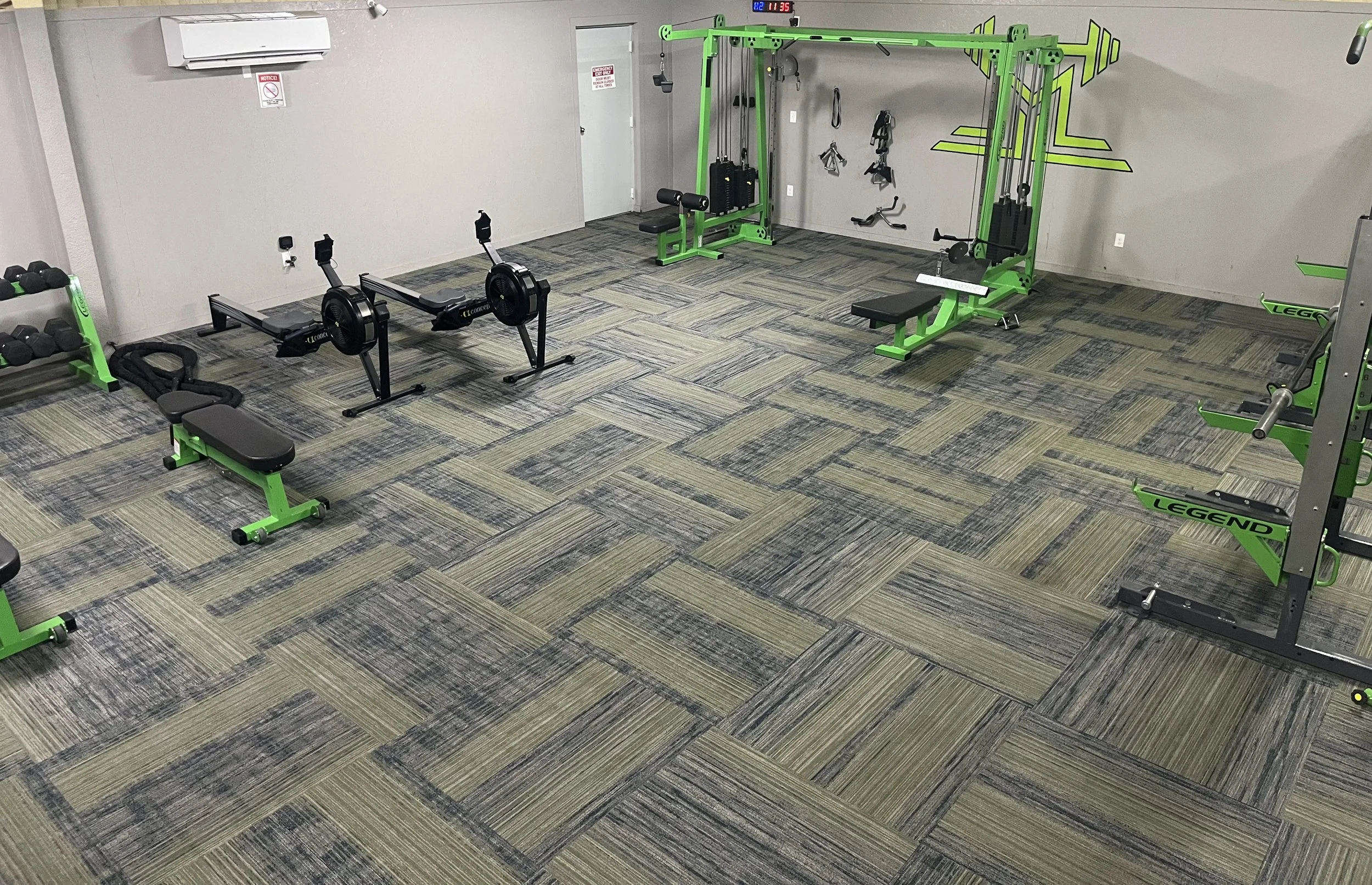 Empty gym with black, green, and gray fitness equipment including rowing machines, weight benches, and a multi-station workout frame.