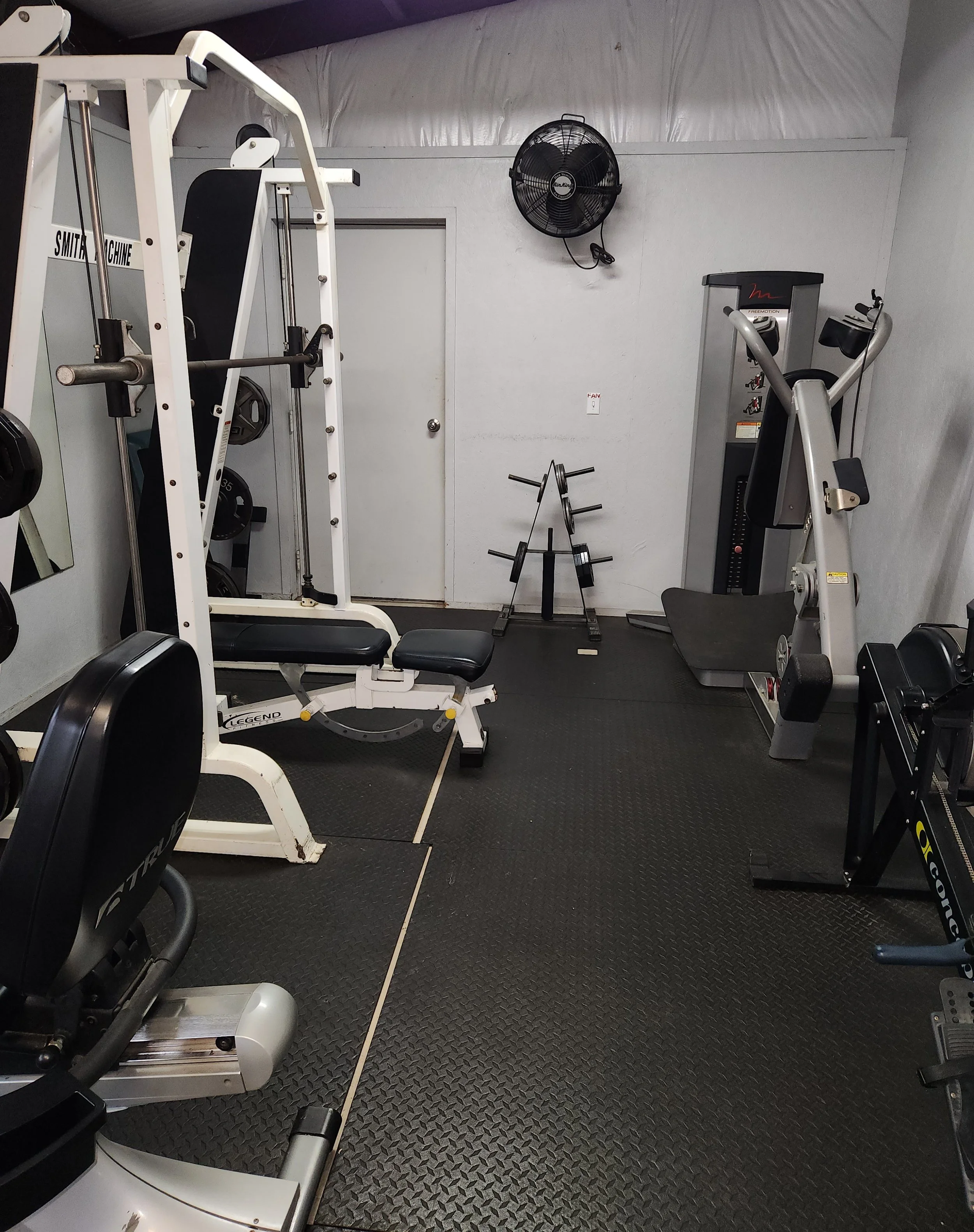 Small gym room with weight equipment including a seated leg press machine, an elliptical trainer, and a pull-down machine, with black rubber flooring and a wall-mounted fan.