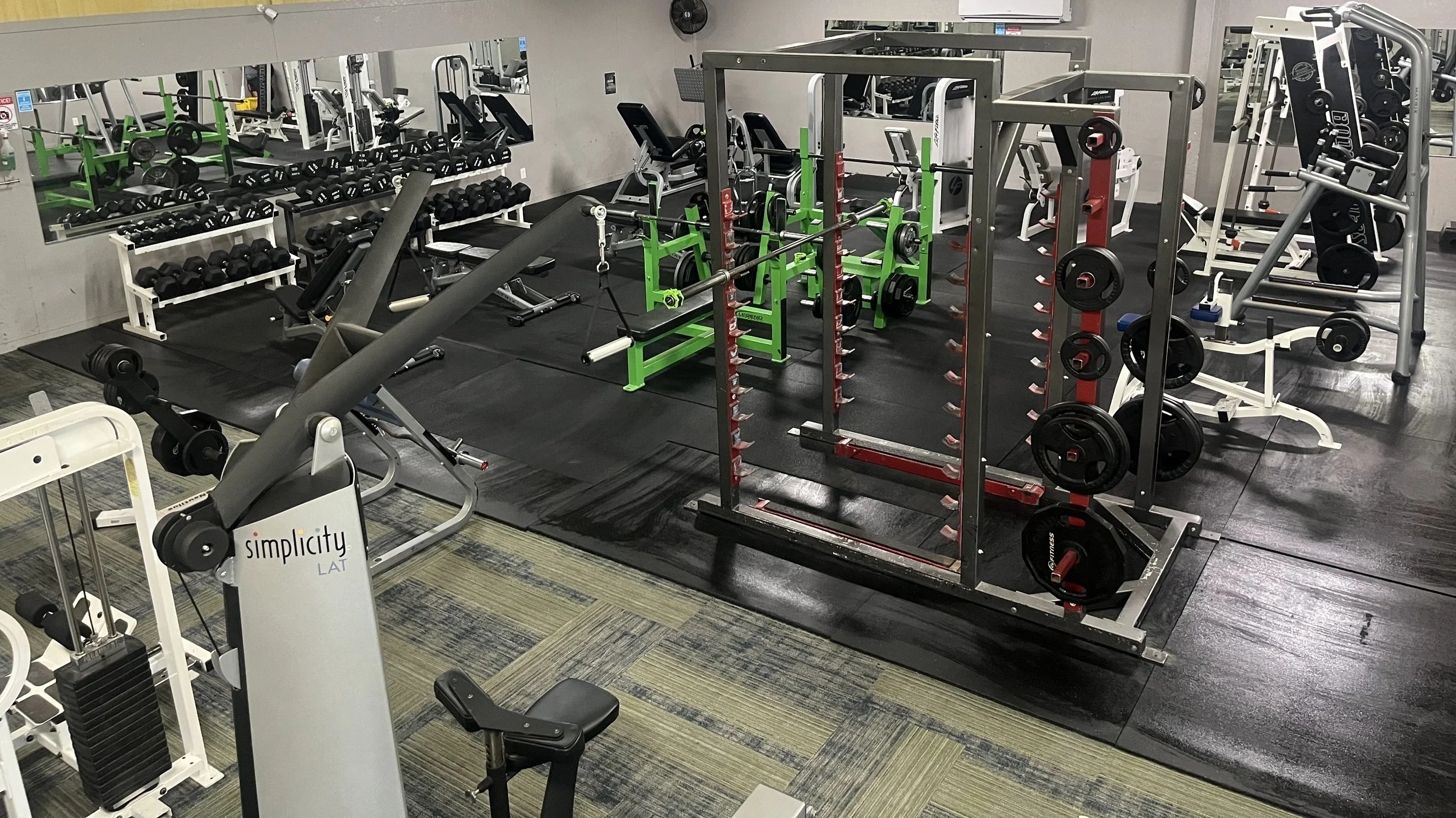 Interior view of a gym with various weightlifting equipment including dumbbells, barbells, squat racks, and benches. The gym has a black rubber floor and mirrors on the wall.