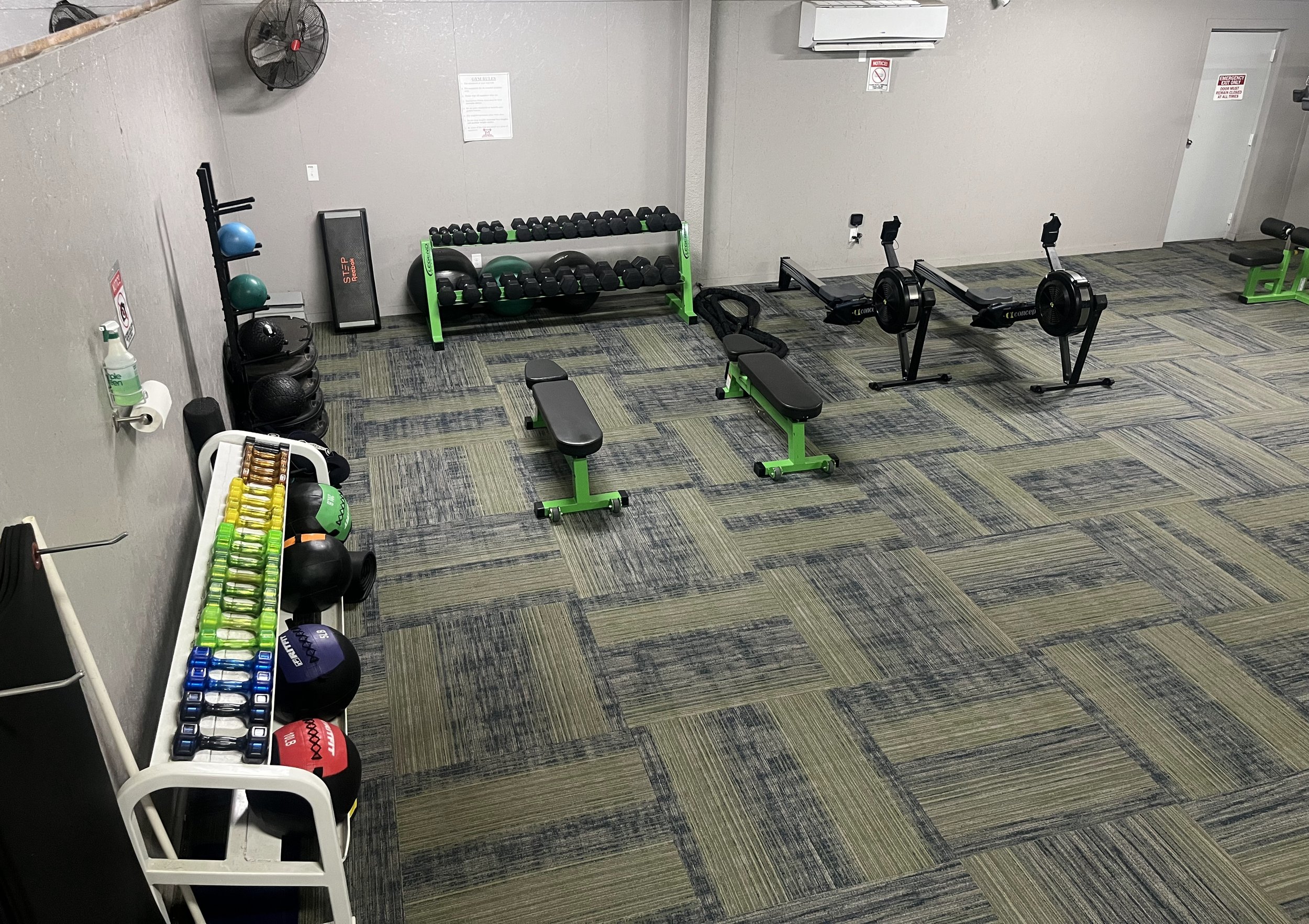 Empty fitness gym with various workout equipment including dumbbells, kettlebells, medicine balls, exercise bikes, benches, and a rack of colorful kettlebells, organized along the walls and on the floor.