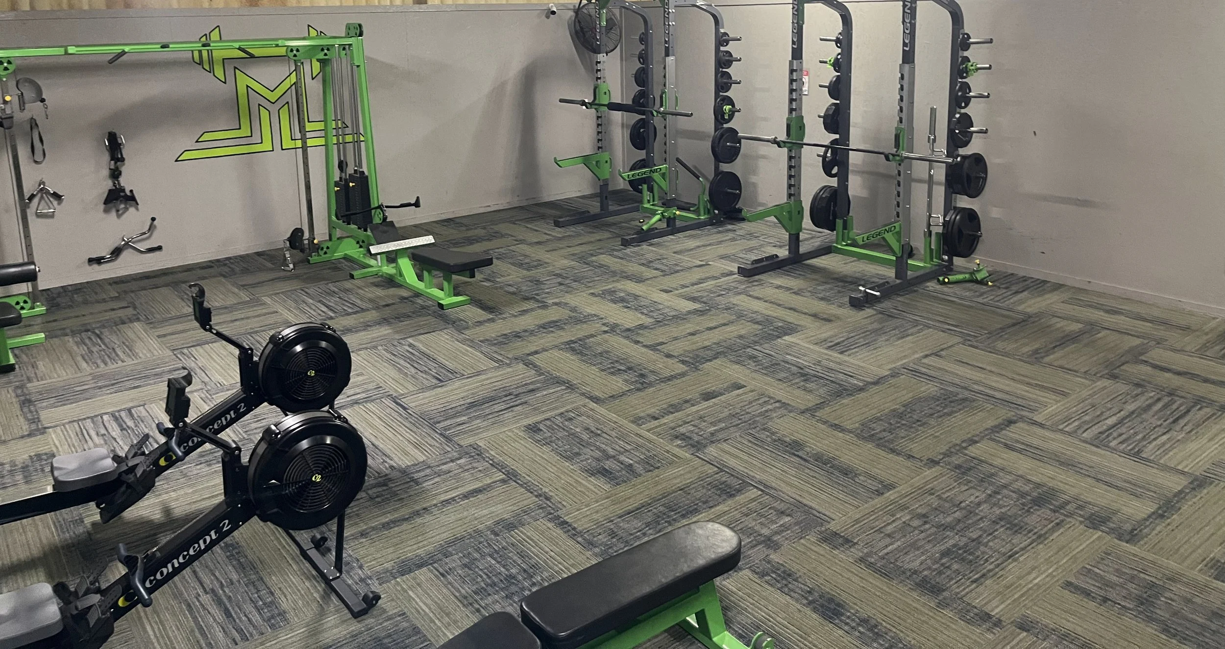 A small gym with various equipment including a rowing machine, bench, and weight racks with plates. The gym has gray and green fitness gear and a patterned carpet floor.
