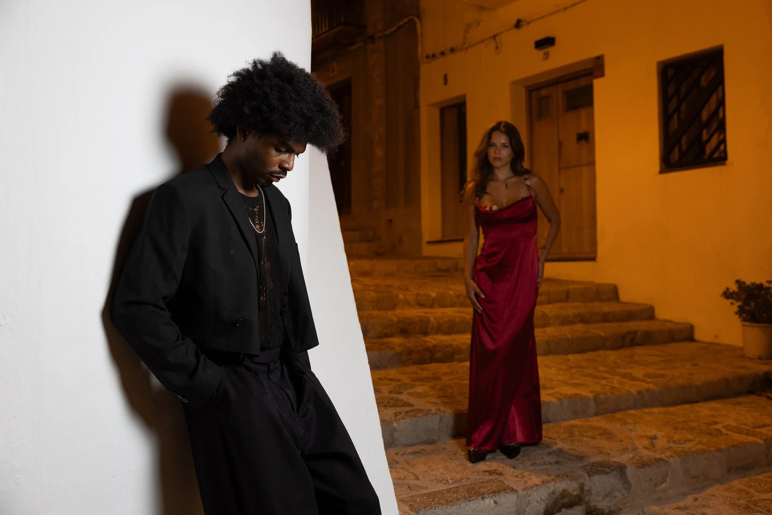 A man with curly hair and a woman in a red dress standing on a stone staircase outside a building at night. The man is leaning against a white wall, looking down, while the woman stands looking at him with her hand resting on her hip.