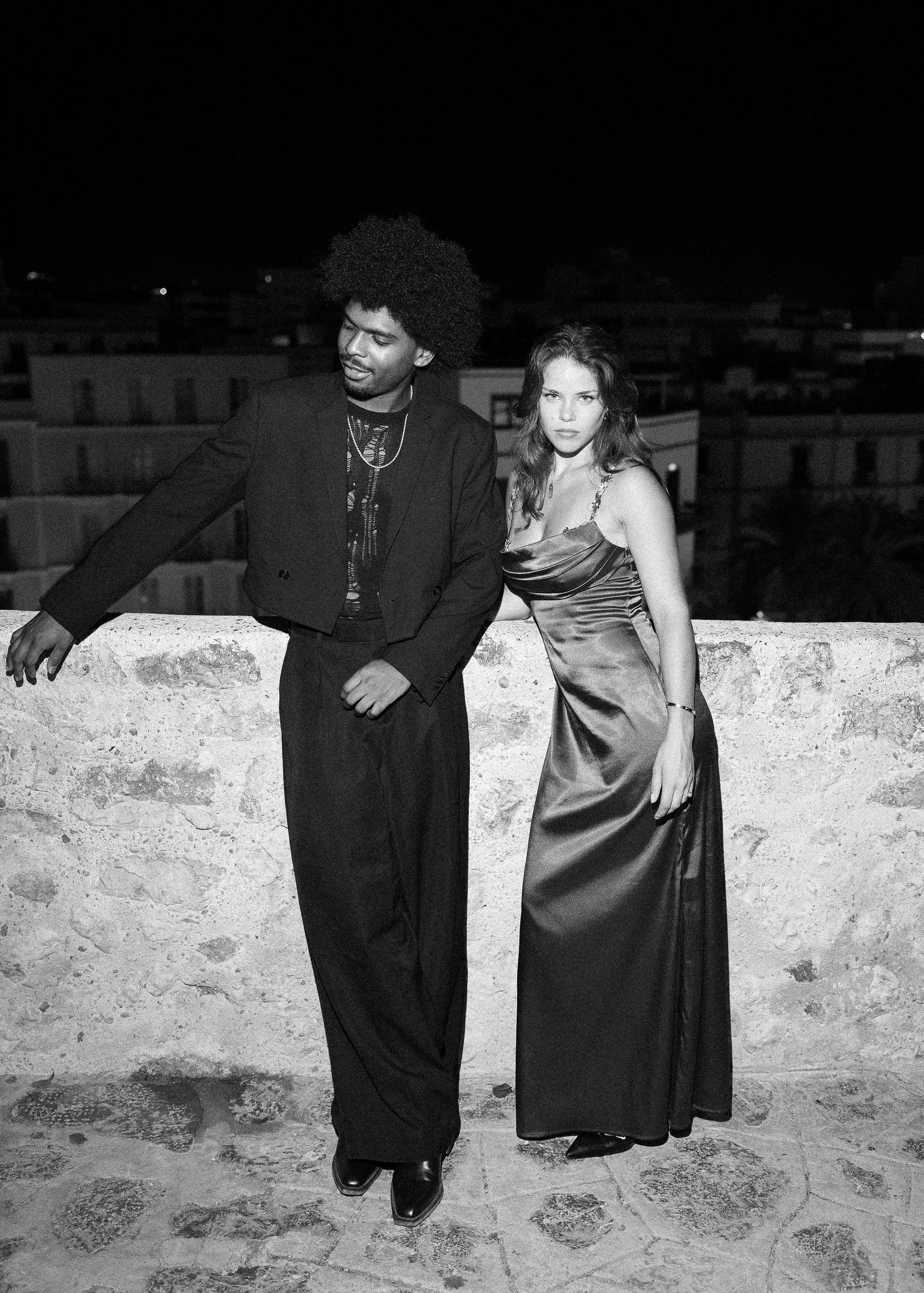 A black-and-white photo of a man and woman dressed in formal attire standing on a rooftop at night, with city buildings in the background.