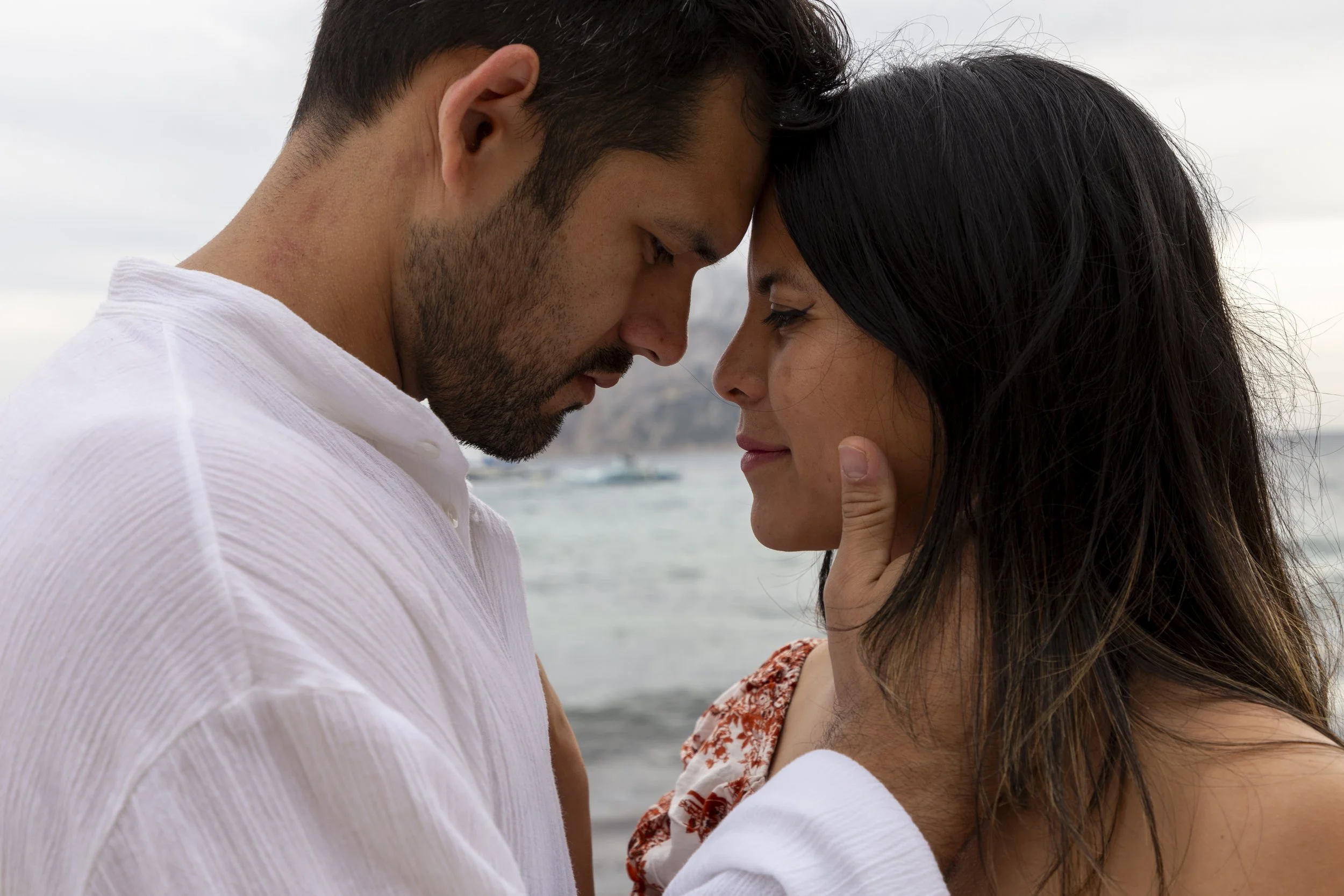 A man and woman with their foreheads touching, gently holding each other's faces, at the beach with the ocean and overcast sky in the background.
