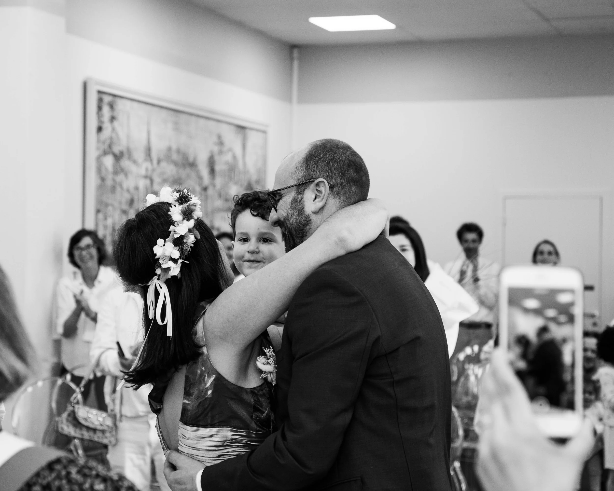 A man in a suit hugging a young girl with flowers in her hair, while holding a young boy who is smiling. Several people are clapping and smiling in the background at an indoor celebration.