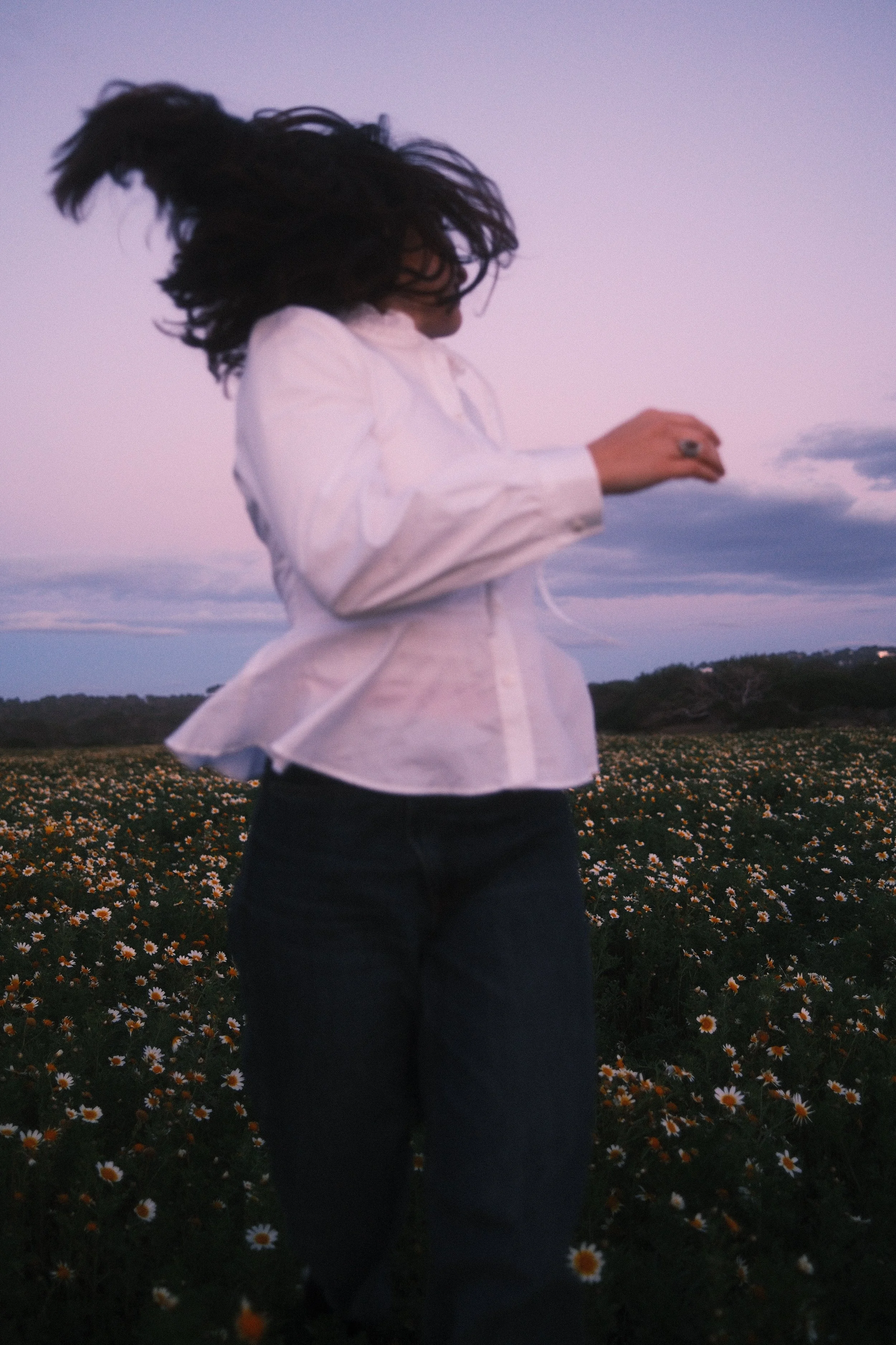 A woman with dark hair in motion, wearing a white shirt and dark jeans, standing in a field of daisies at dusk.