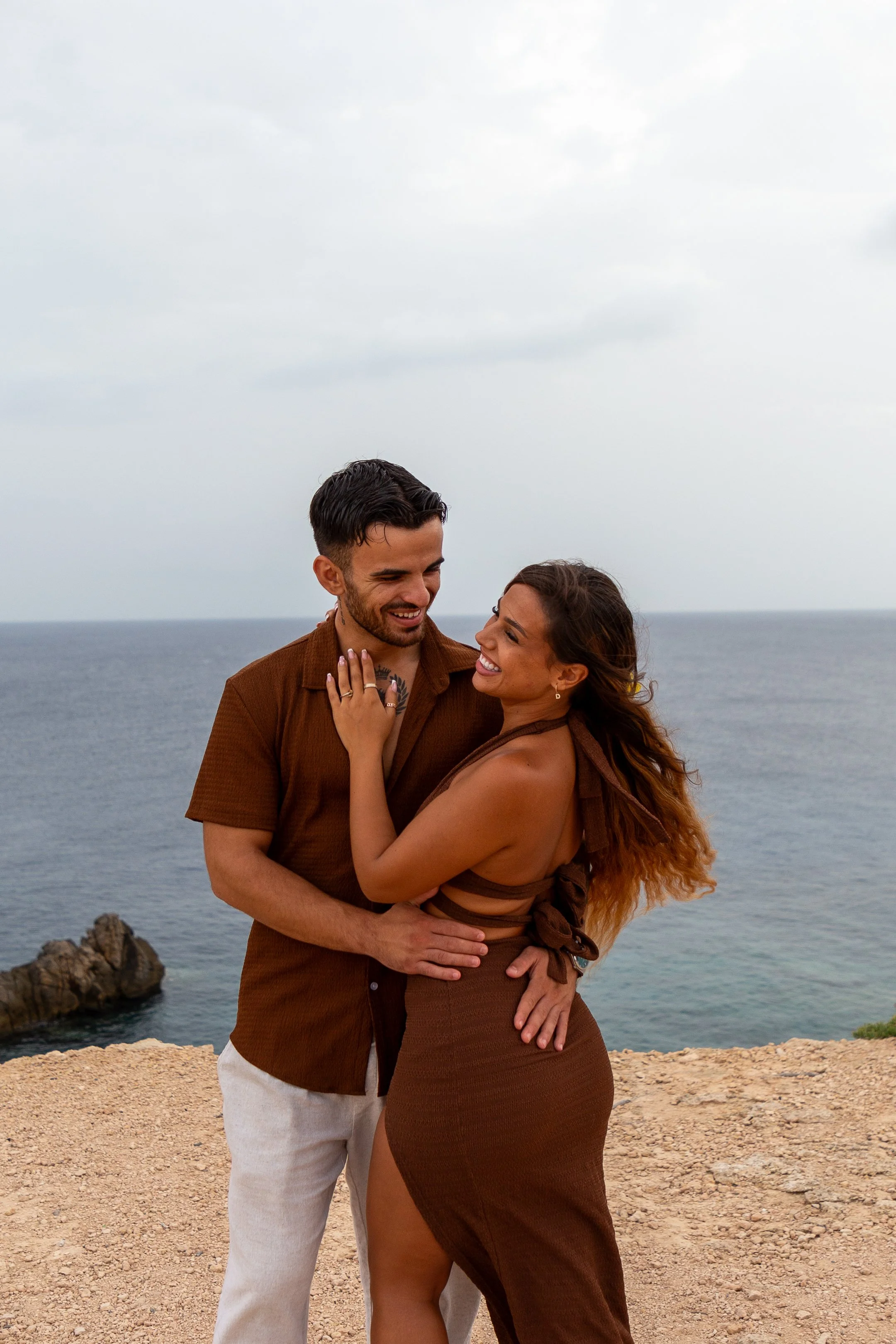 A smiling couple embracing each other on a rocky cliff by the sea, with an overcast sky overhead.